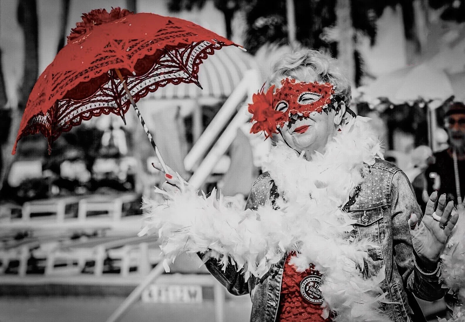 A woman wearing a red masquerade mask and red lipstick, holding a red lace parasol, dressed in a feather boa, and wearing a badge. The background appears to be an outdoor setting with blurred figures and objects, mostly in black-and-white.