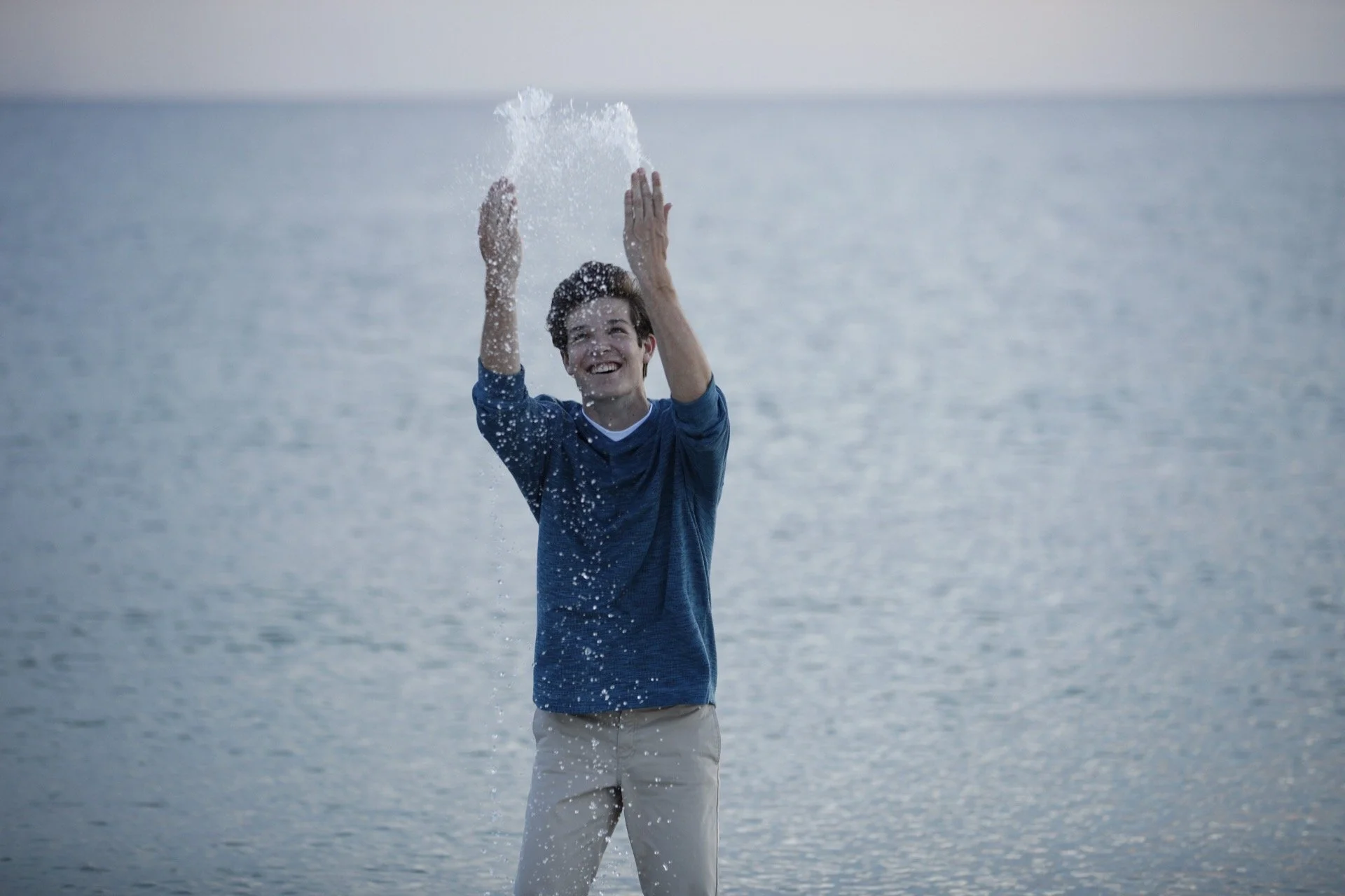 A young man in a blue long-sleeve shirt and khaki pants is standing by a body of water, smiling and tossing water into the air, with a gray sky and calm water in the background.
