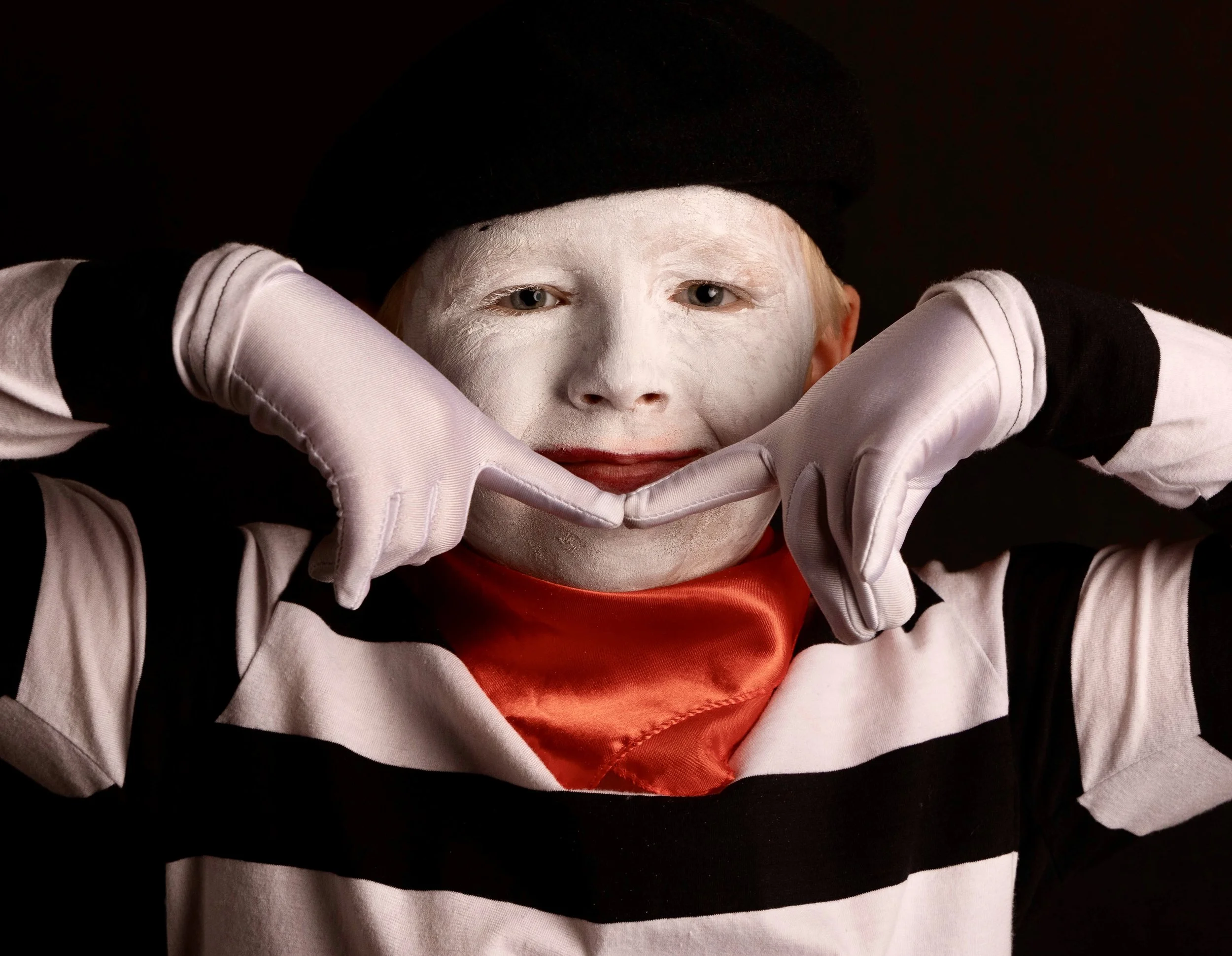 A person with white face paint, red lipstick, and blonde hair wearing a black beanie and black-and-white striped shirt, holding hands in front of their face in a framing gesture.