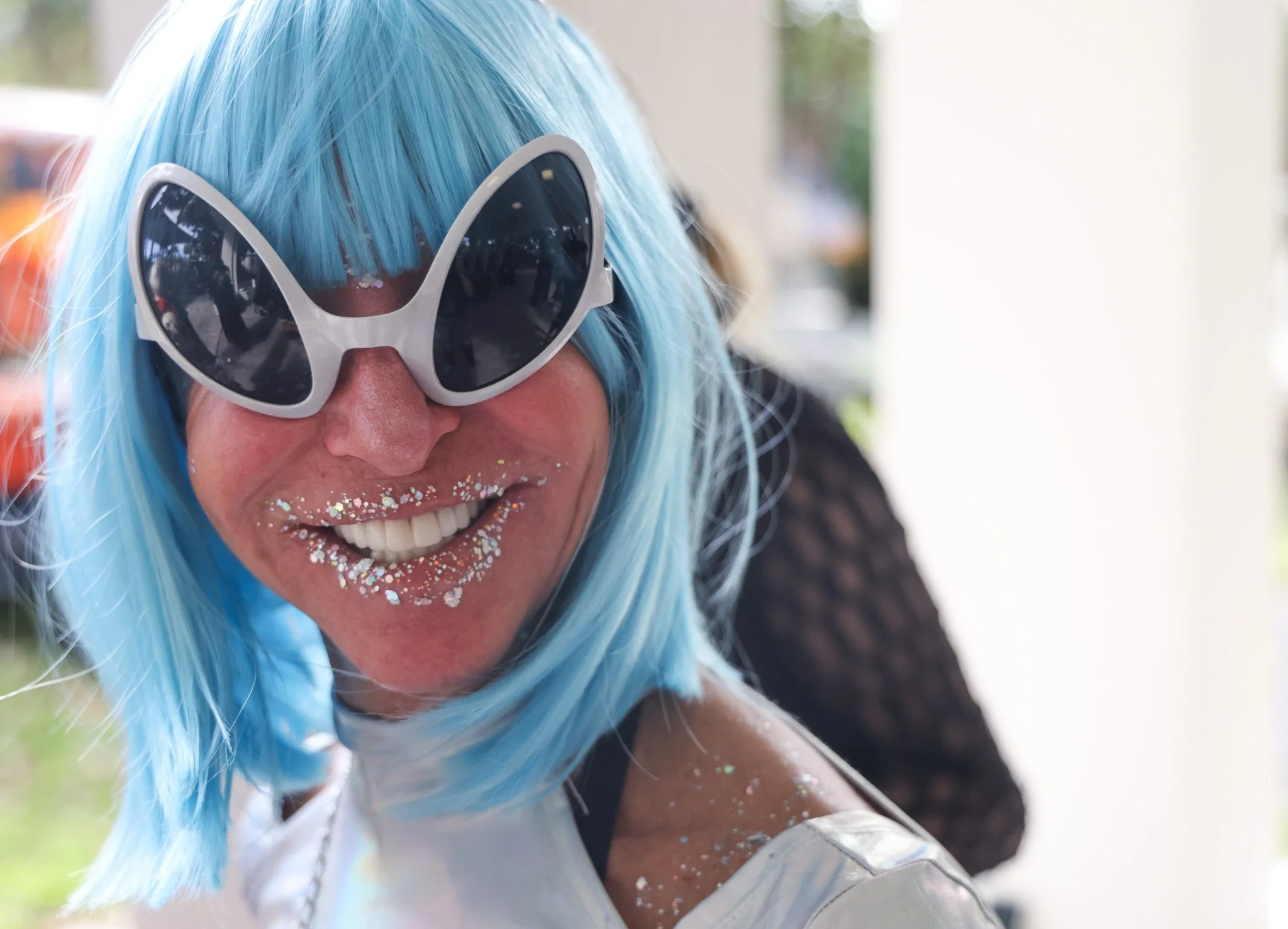 Person with blue hair wearing large sunglasses smiling with glitter on their face and looking at the camera.