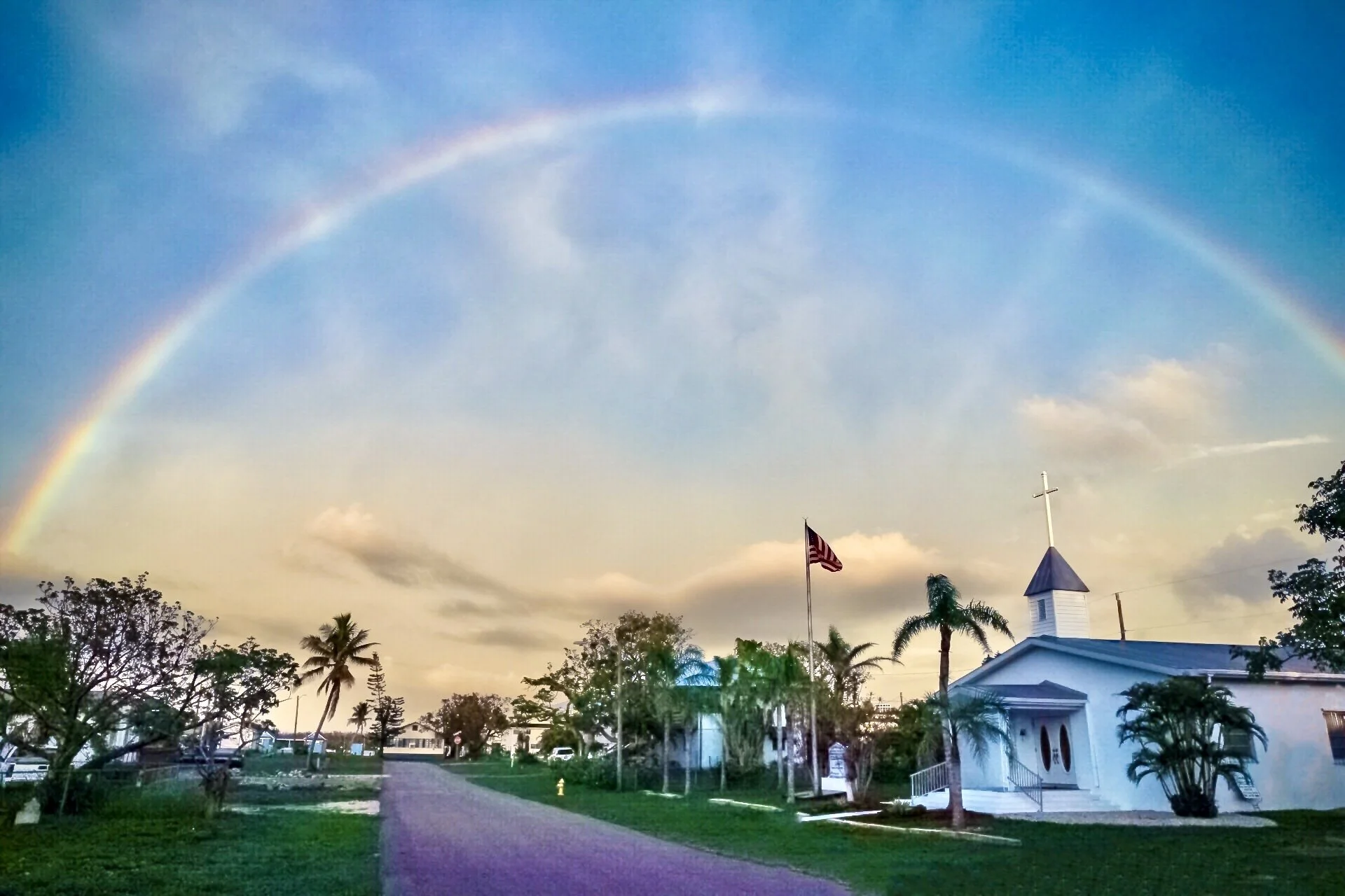 A rainbow arching across a blue sky with some clouds. In the foreground, there are trees, a sidewalk, a small white church with a steeple, and a flagpole with an American flag.