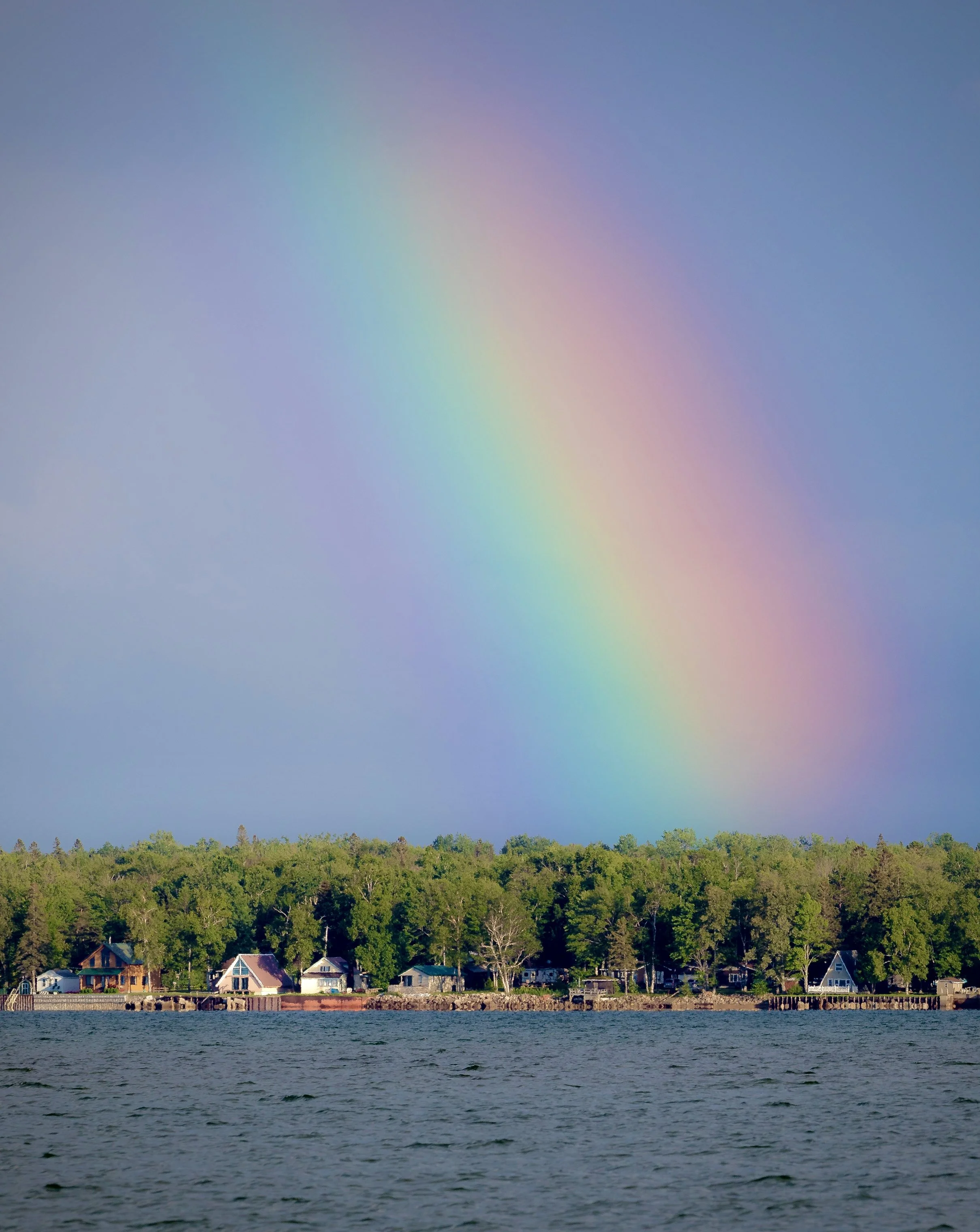 A rainbow arching over a small lakeside community with houses, trees, and a body of water in the foreground.