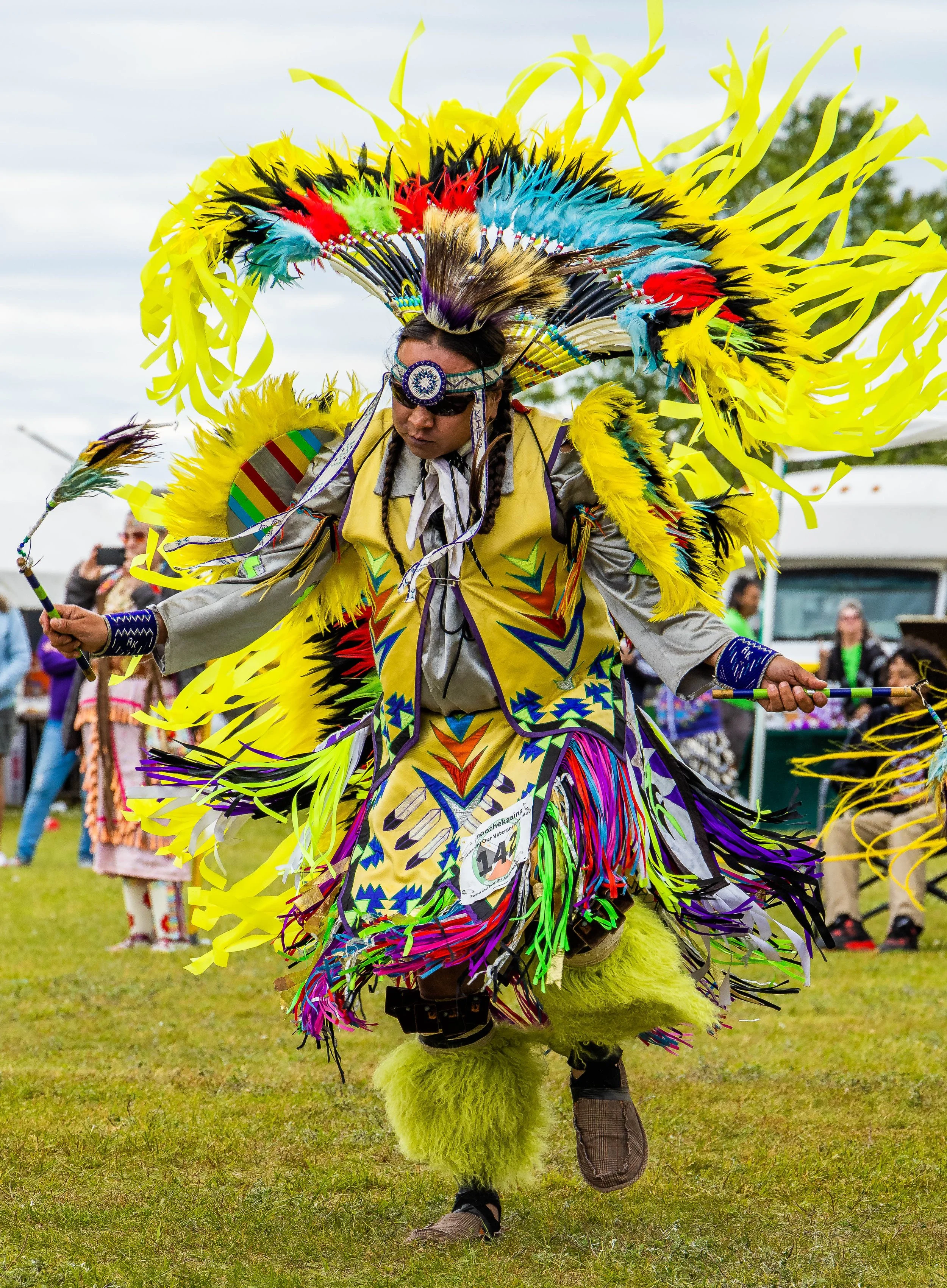 A performer dressed in a vibrant, elaborate costume with colorful feathers, fringe, and traditional patterns, participating in a cultural festival or parade outdoors, surrounded by onlookers.