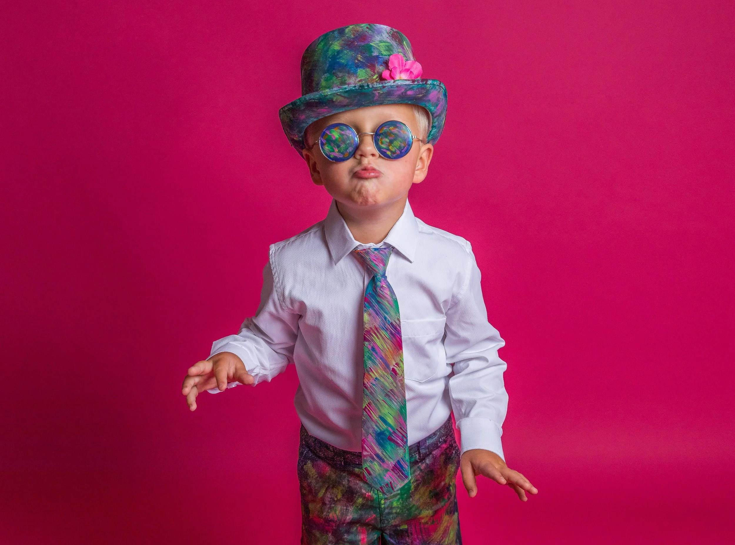 A young boy in colorful, abstract-patterned clothing and accessories against a pink background, wearing round, rainbow-colored sunglasses and a matching colorful hat with a pink flower, with a confident pose.