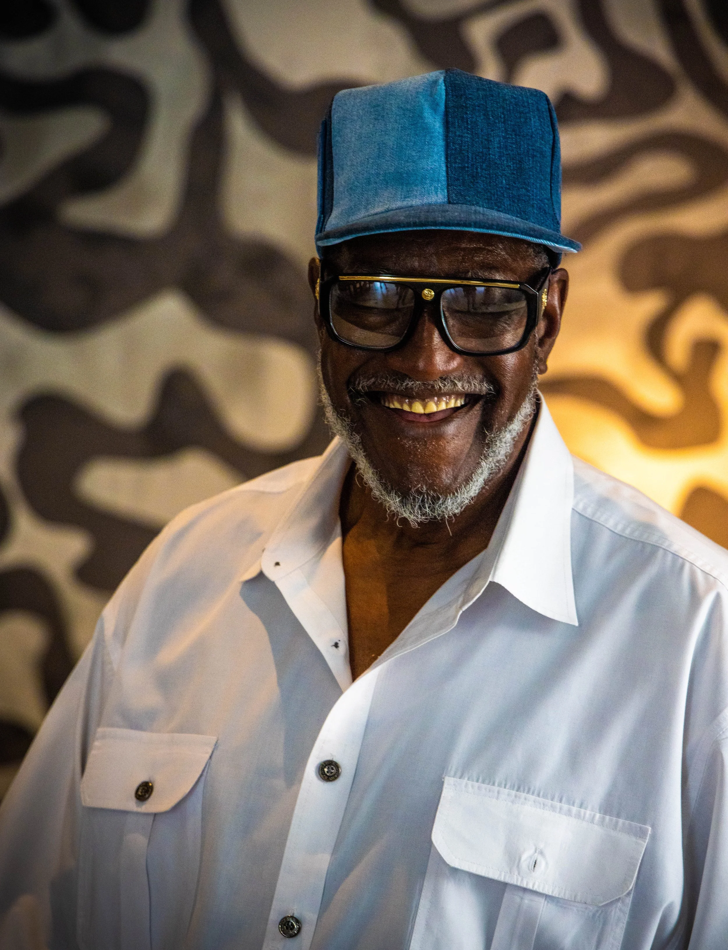 A smiling man wearing a blue baseball cap, black-rimmed glasses, and a white button-down shirt standing in front of a patterned background.
