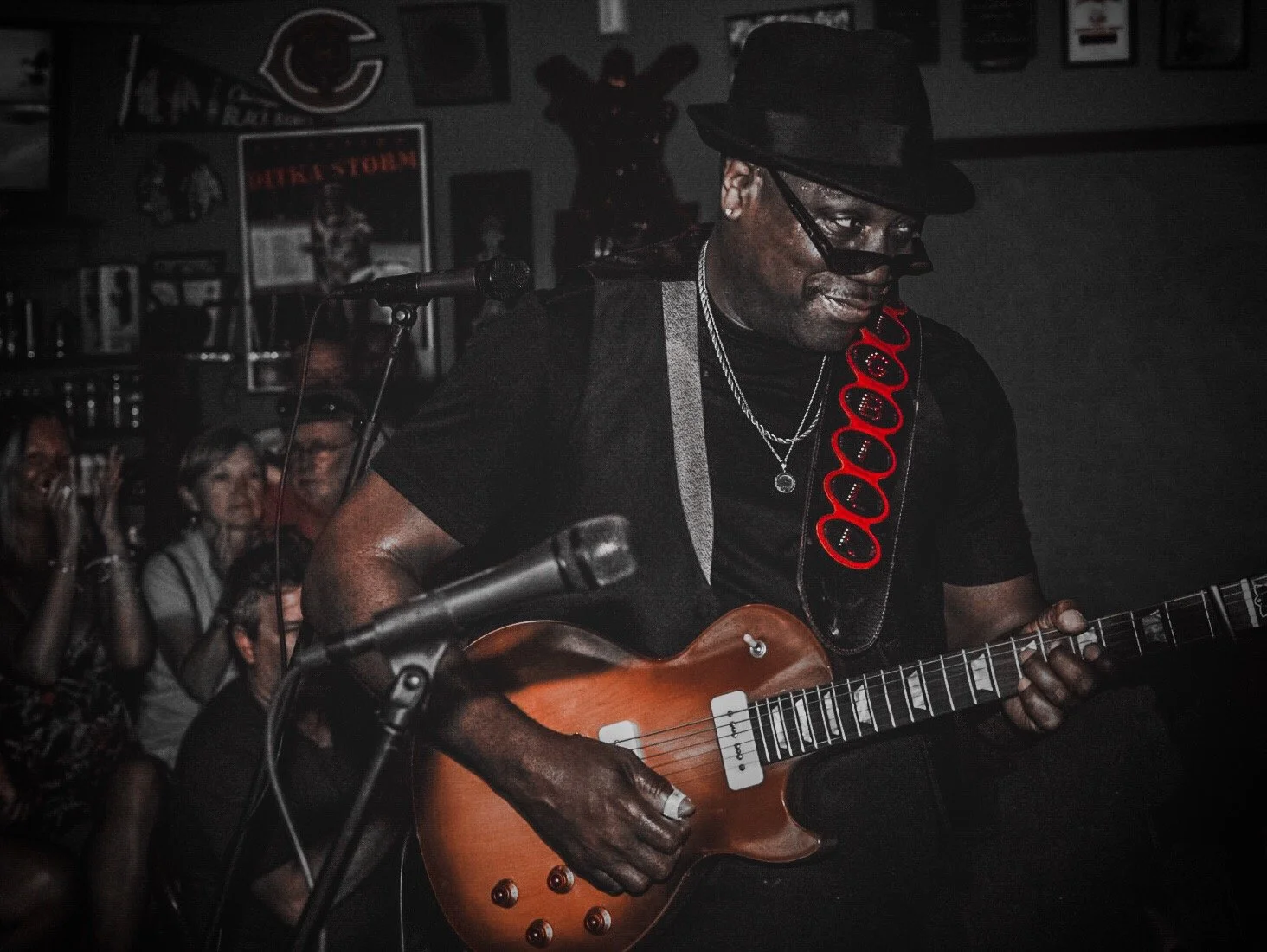 A man playing an electric guitar in a dimly lit room with an audience in the background.