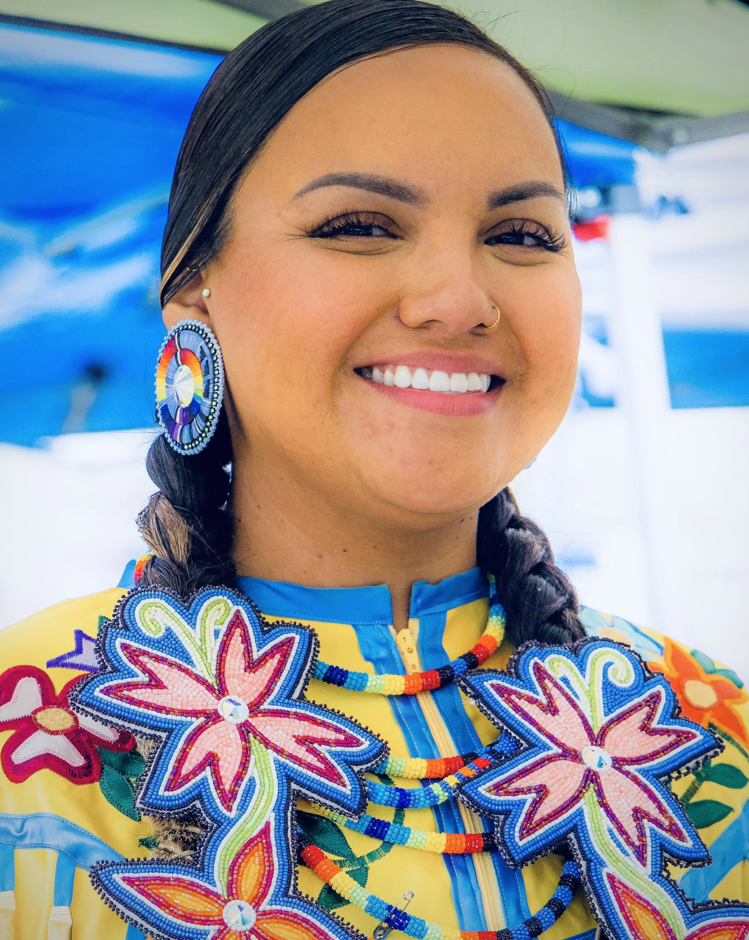 A woman smiling, wearing colorful traditional Indigenous clothing and jewelry, with beadwork designs including flowers and geometric patterns, jewelry featuring large beaded earrings and necklaces, and her hair styled in braids.