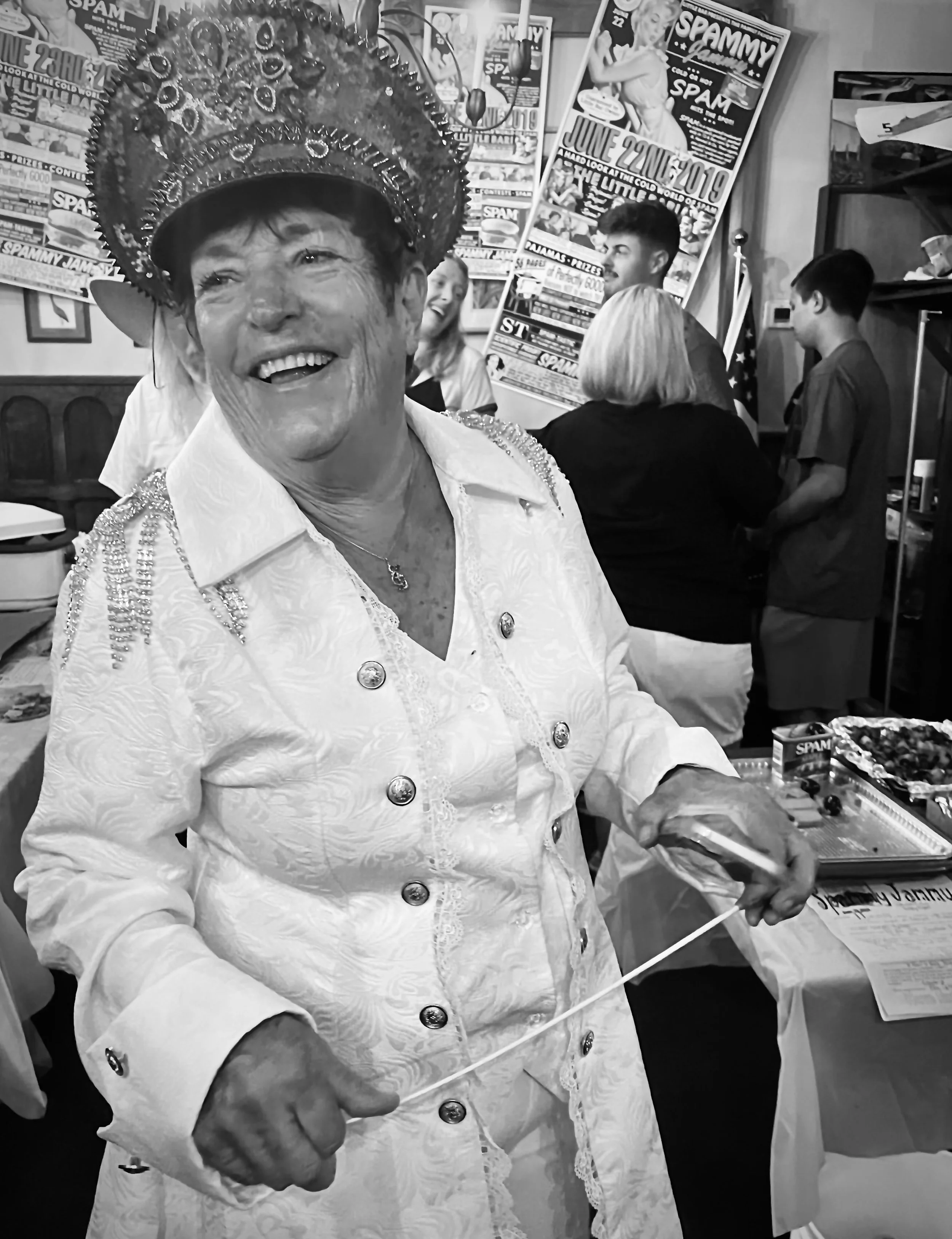 An elderly woman wearing an ornate crown and a white embroidered jacket, smiling happily, standing at a table with a box of Spam and other items. A group of people and posters are visible in the background at an event or gathering.