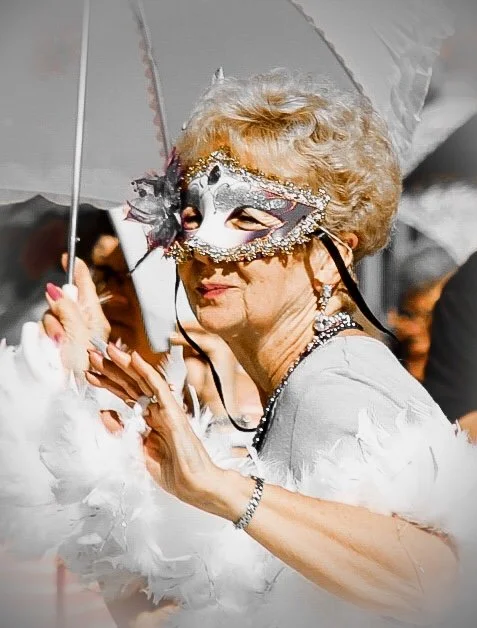 An elderly woman in a fancy dress and jewelry wearing a silver mask with black feathers, holding an umbrella at a festive outdoor event.