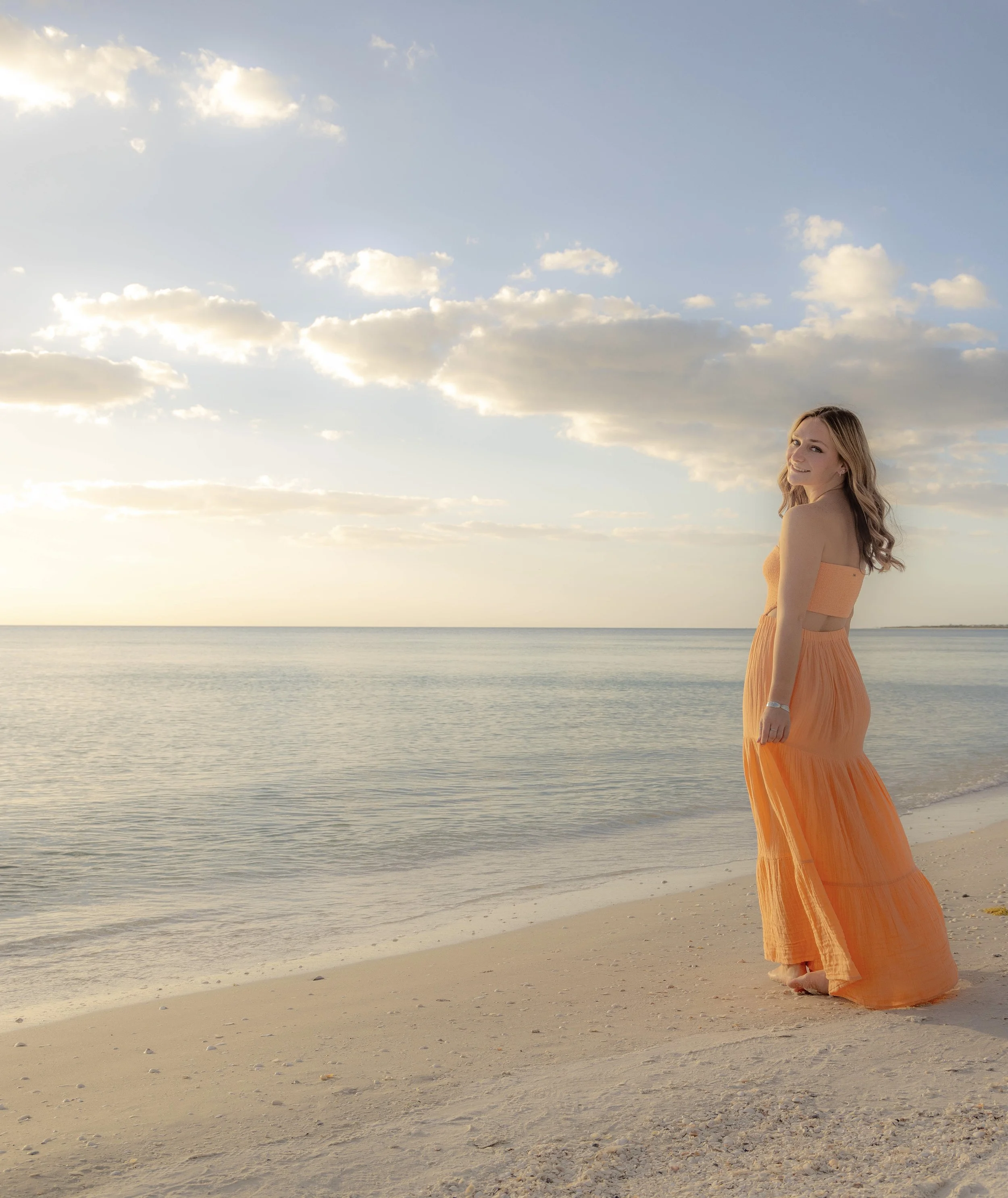 A woman in an orange dress standing barefoot on a sandy beach, looking back and smiling with a partly cloudy sky and calm ocean behind her