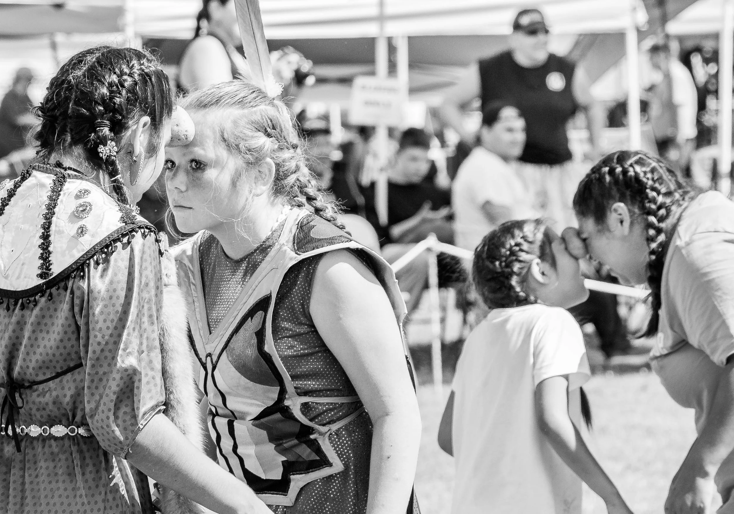 Two girls, one with braided hair and the other with curly hair and freckles, face each other closely at an outdoor event, with other people and tents visible in the background.