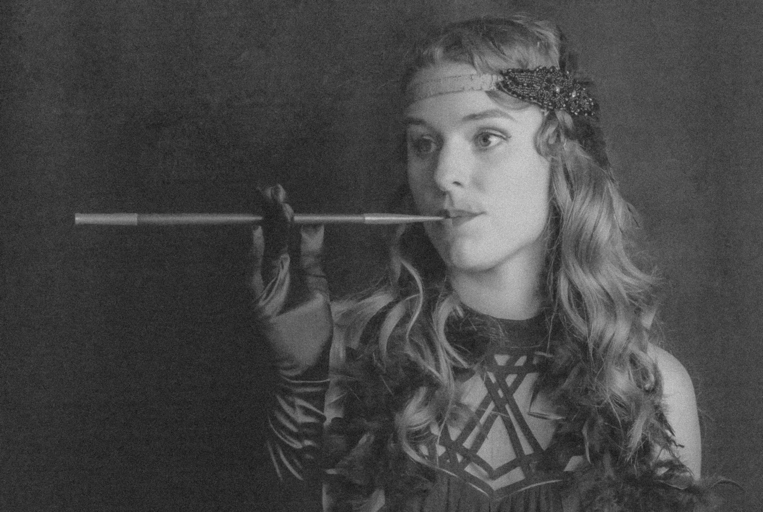 A woman with curly hair and a headband, holding a smoking cigarette in her hand, looking to the side with a neutral expression.