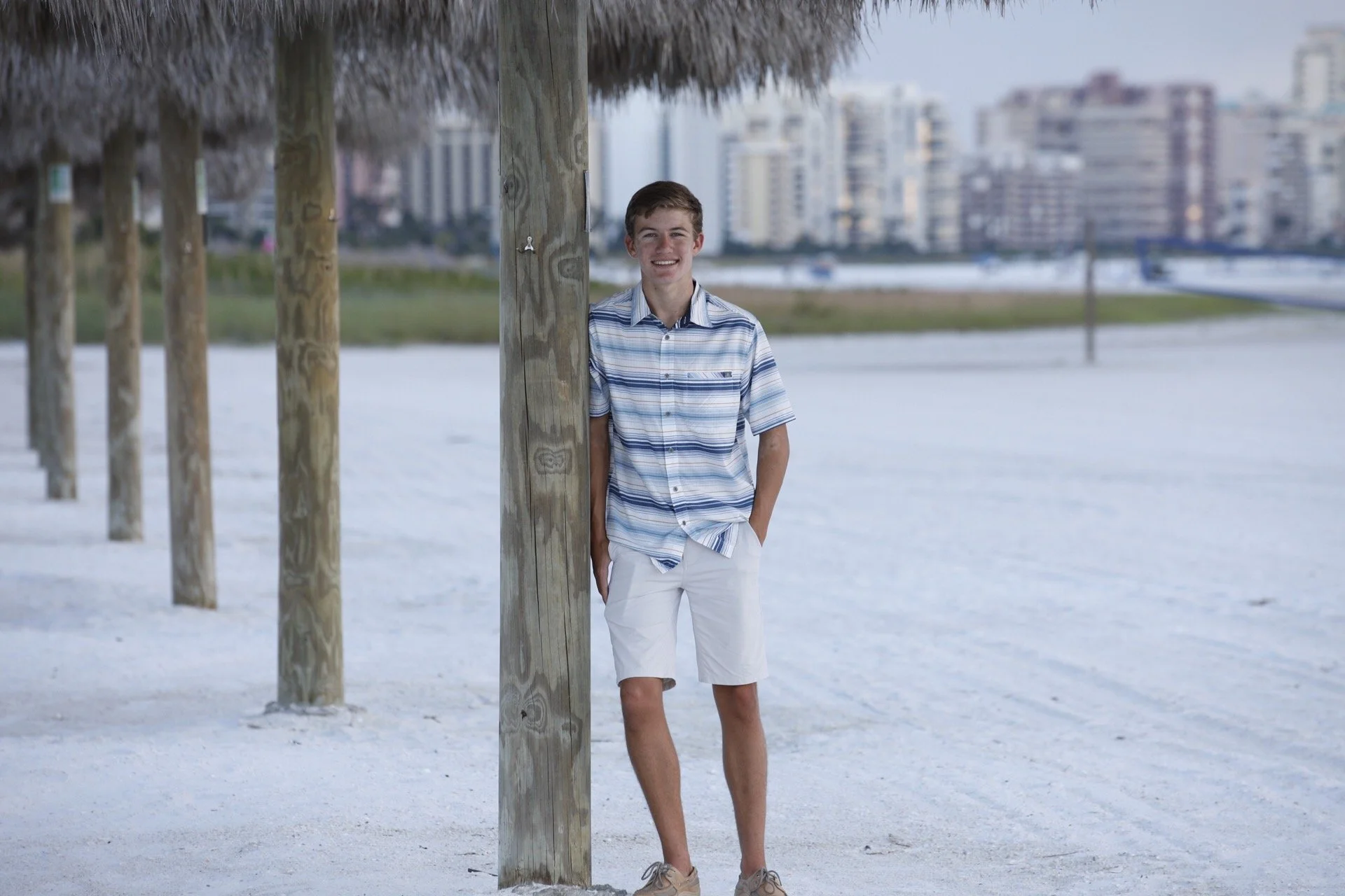 A young man standing on a white sandy beach, leaning against a wooden pole with a thatched roof, smiling at the camera with city buildings in the background.