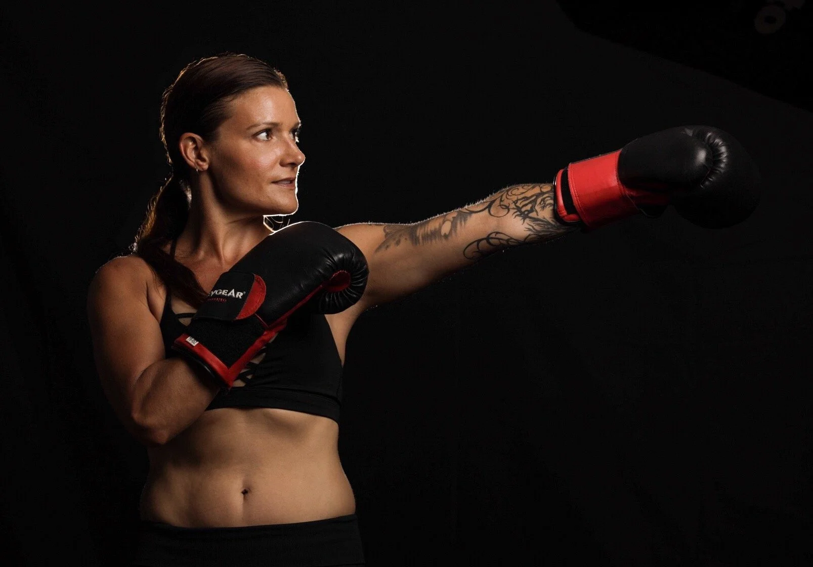 A woman in black sports bra and boxing gloves throws a punch against a black background.