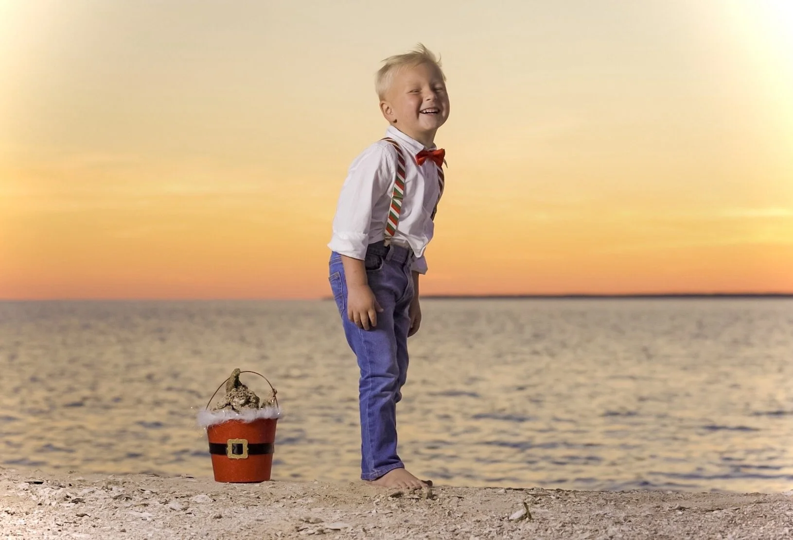 A young boy in a white shirt, red bow tie, suspenders, and jeans stands barefoot on a beach at sunset, smiling and looking to the side. There is a small pirate hat with a feather and a decorative buckle on the ground nearby.