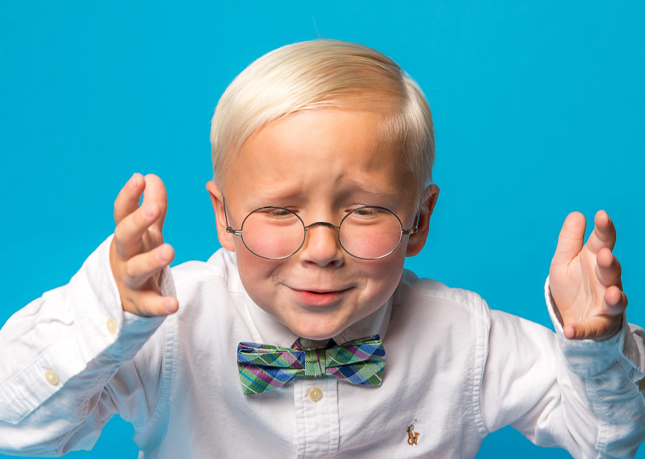 A young boy with blonde hair and glasses wearing a white shirt and a colorful bow tie, smiling and making a playful gesture with his hands against a blue background.