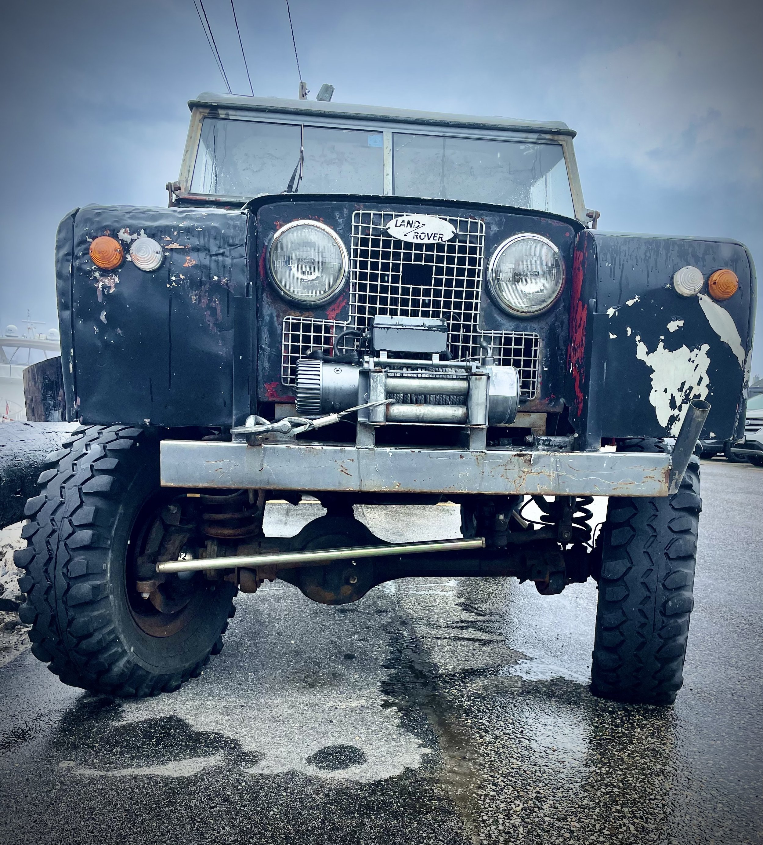 Close-up front view of a vintage black Land Rover with large off-road tires, exposed mechanical parts, and a weathered appearance, parked on a wet asphalt surface.