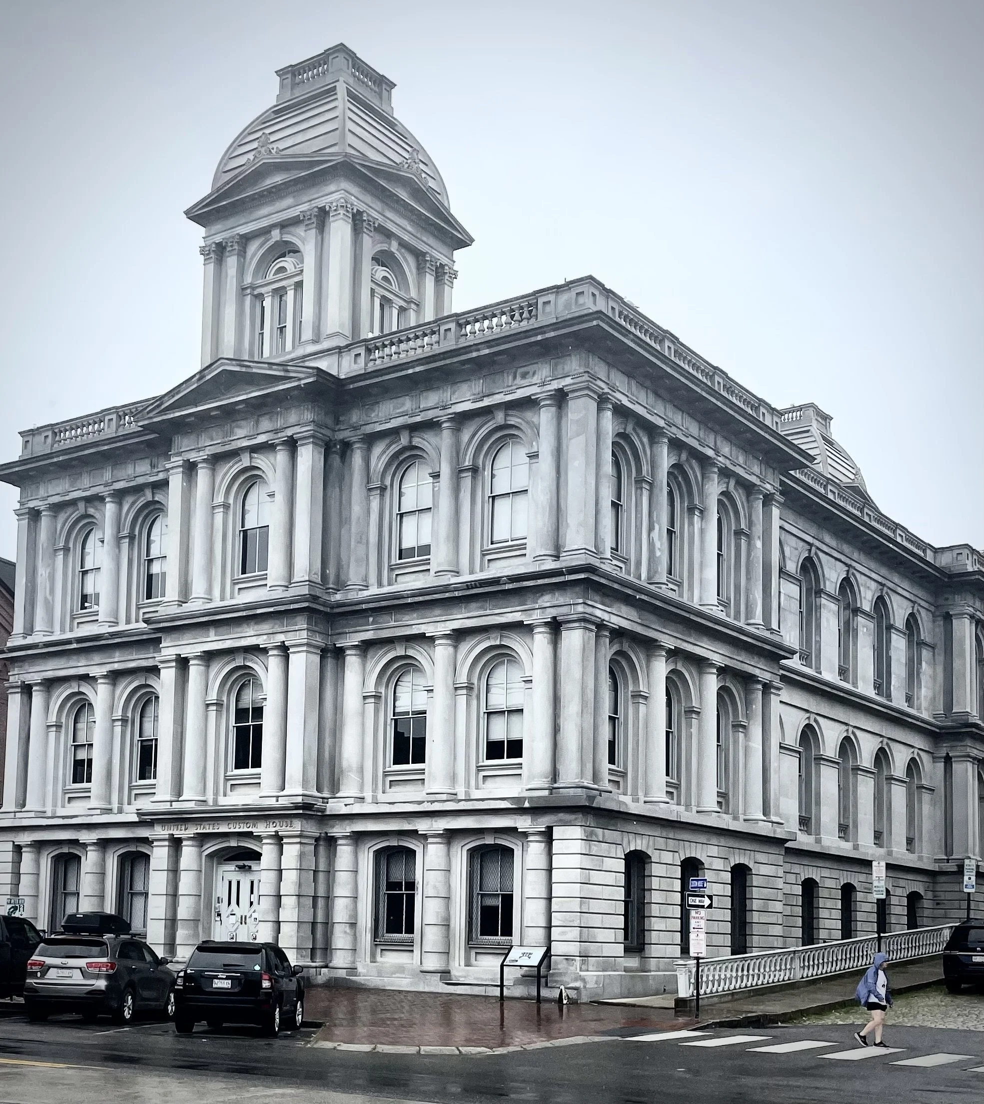 Historic courthouse building with classical architecture, featuring columns and arched windows, and a dome on top. Cars are parked in front, and pedestrians are walking on the sidewalk.