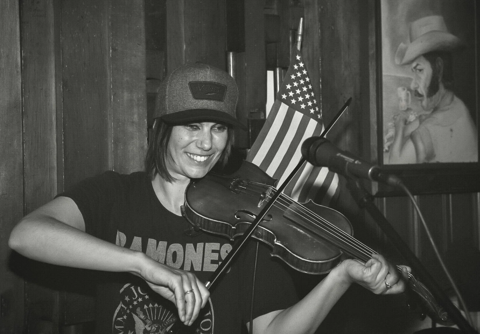 A woman playing the violin, smiling, wearing a baseball cap and a Ramones t-shirt, with an American flag behind her, and a portrait of a woman with a cowboy hat on the wall.