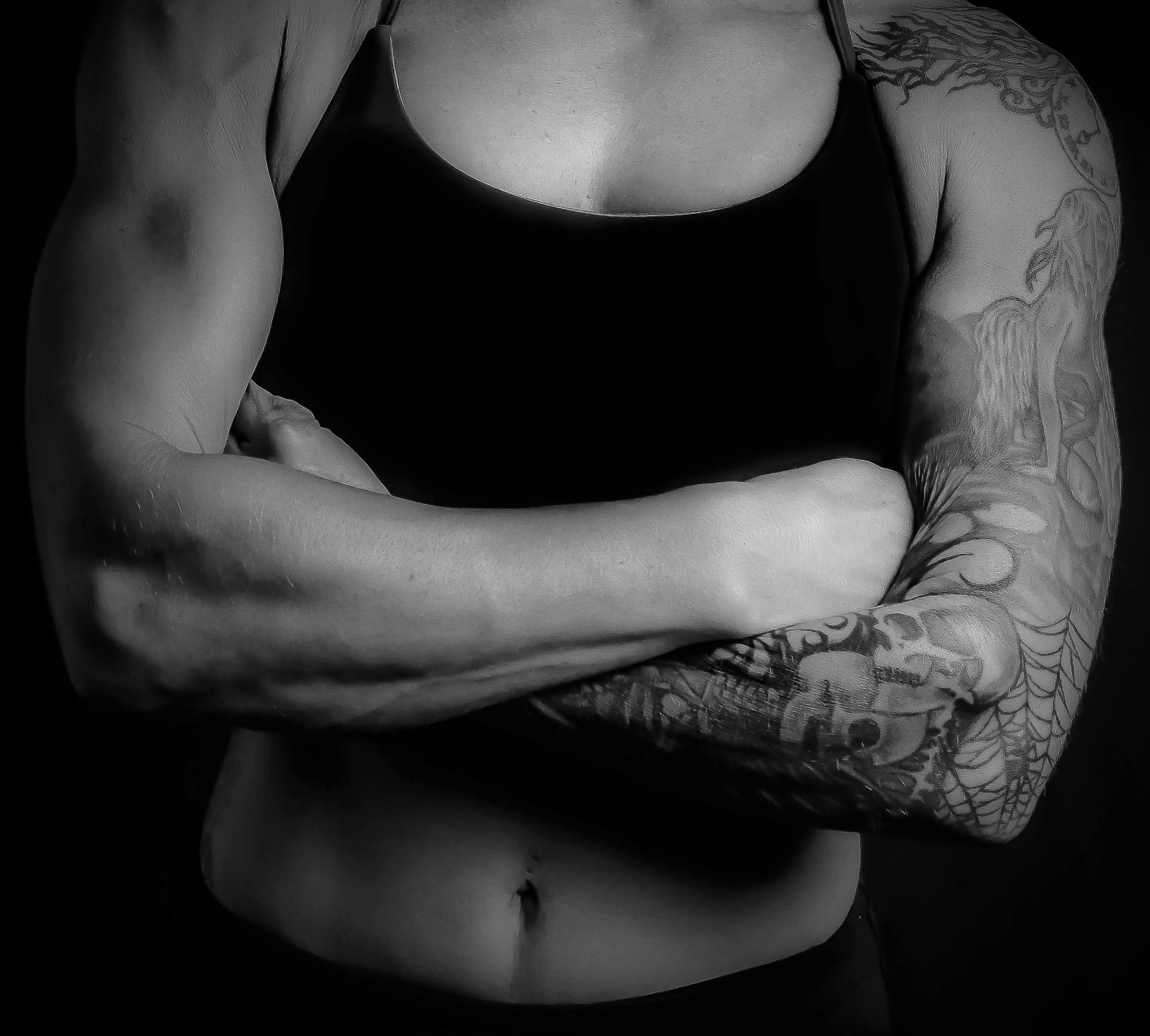 Black and white photo of a person's torso and arm with tattoos, wearing a sleeveless top, crossing arms. The person's face is not visible.