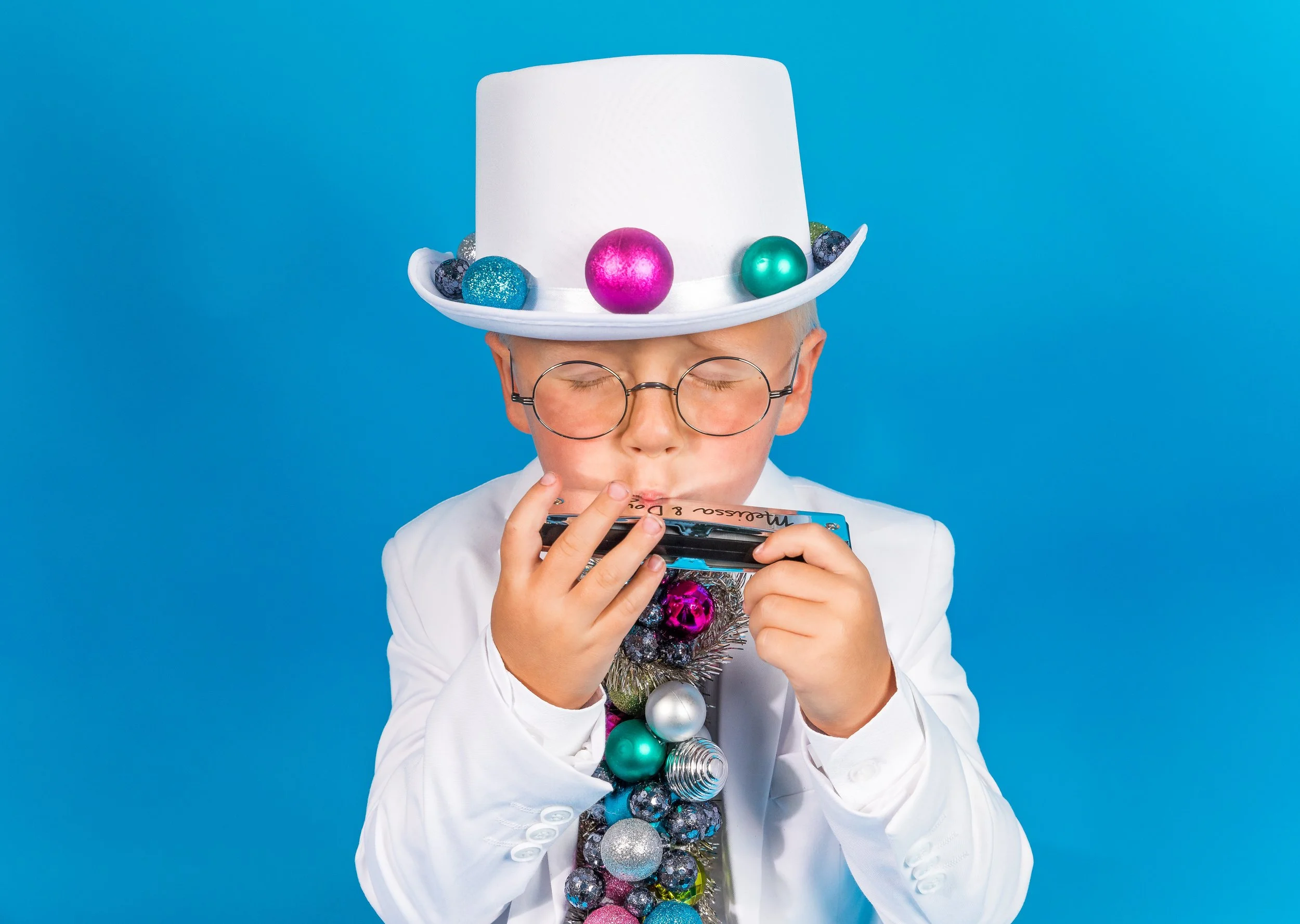 A child wearing glasses and a large white top hat decorated with colorful shiny Christmas ornaments, against a blue background.