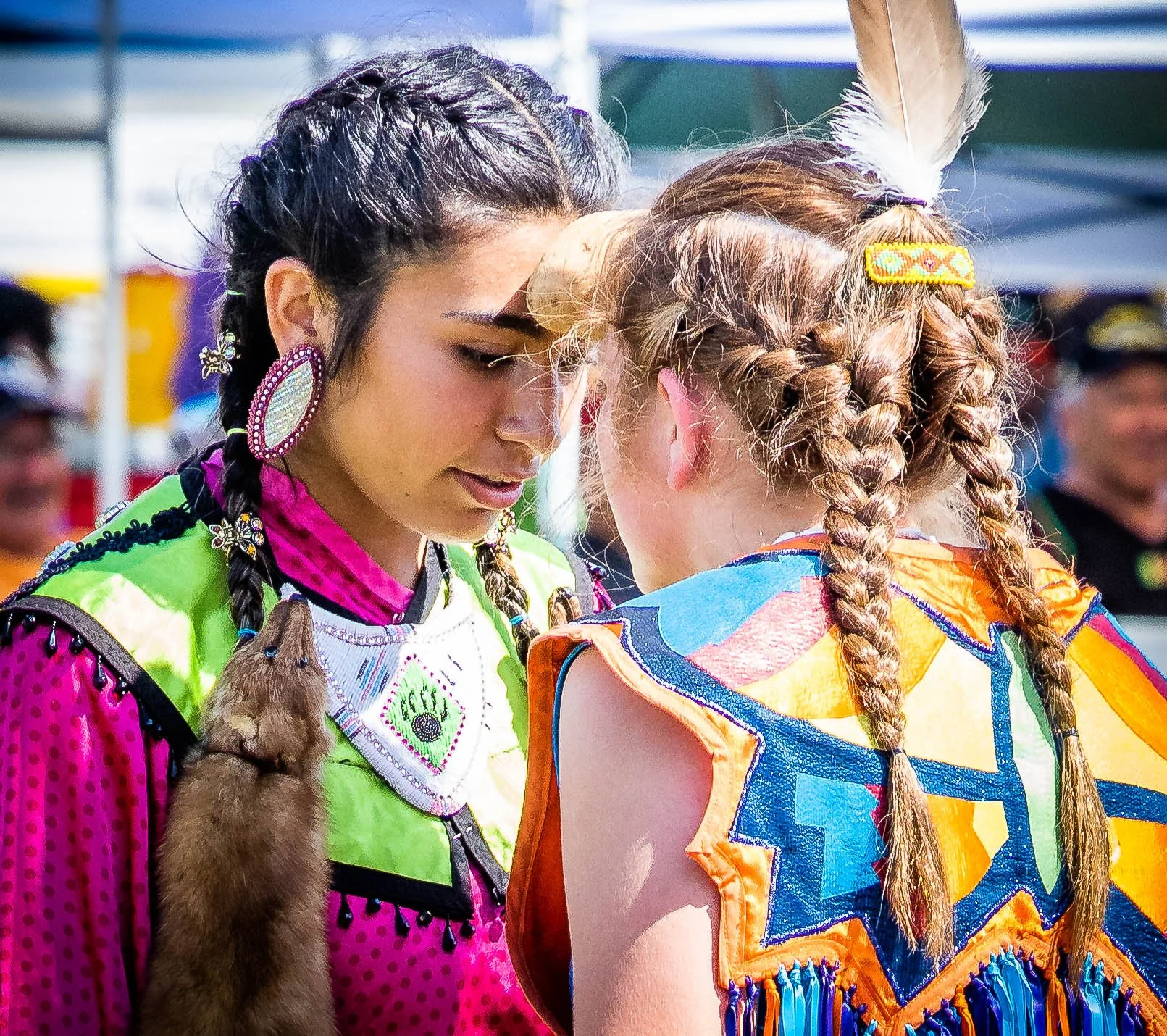 Two Indigenous women face-to-face, touching foreheads, dressed in colorful traditional attire and hairstyles, with one wearing feathers and beaded jewelry, outdoors with a blurred crowd in the background.
