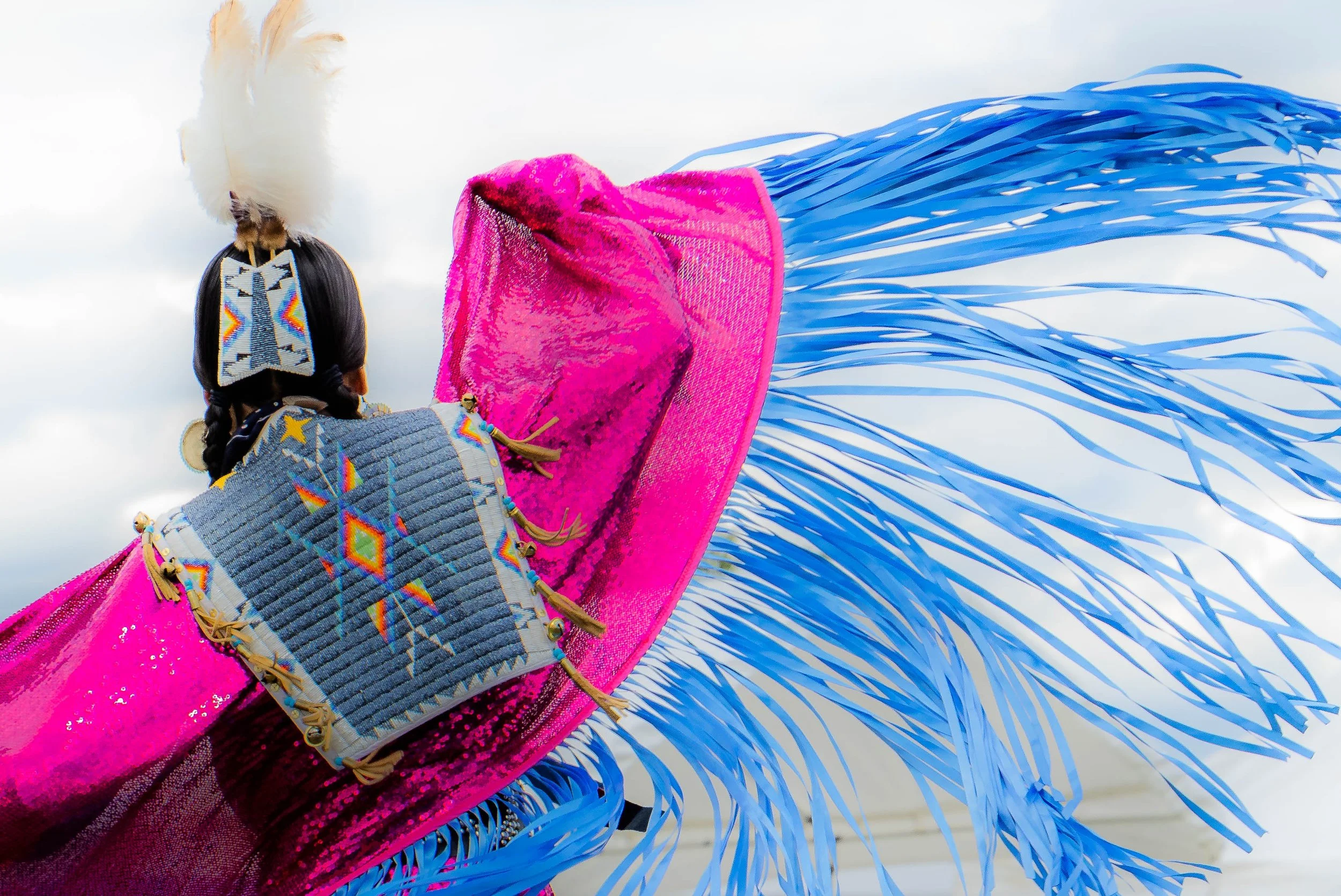 Close-up of a dancer's costume with bright pink fabric, fringes, and beaded details, set against a cloudy sky.