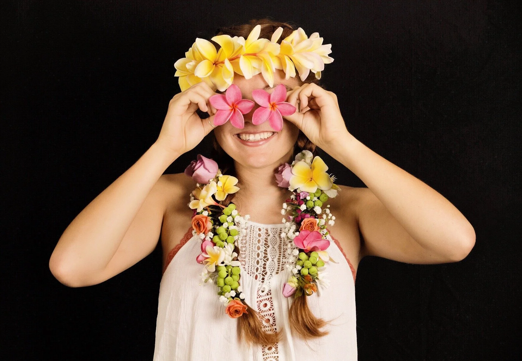 Woman smiling wearing a floral lei and a flower crown covering her eyes, set against a black background.