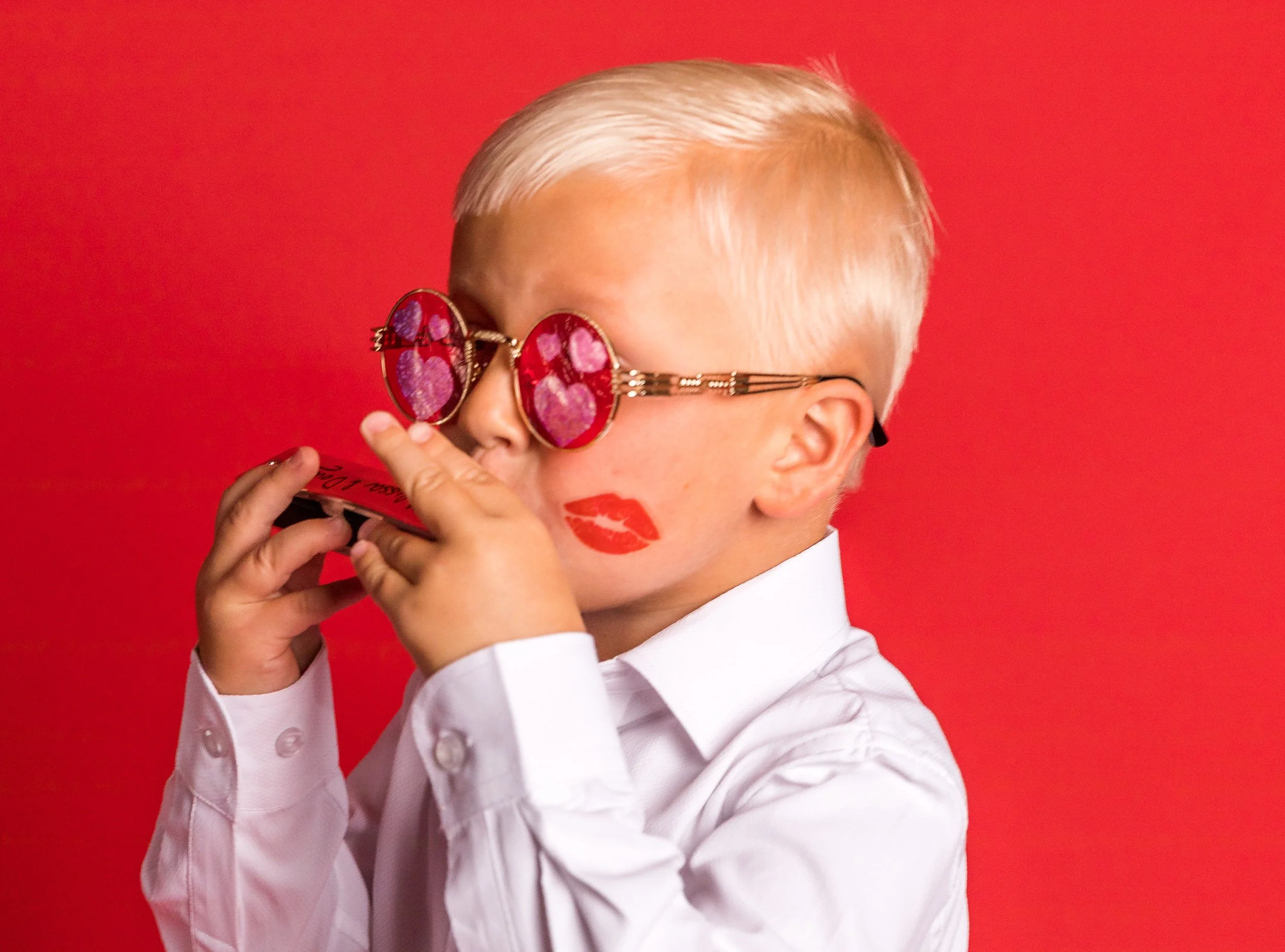 Person with platinum blonde hair wearing heart-shaped sunglasses with pink lenses, holding a red lipstick, and wearing a white shirt against a red background.