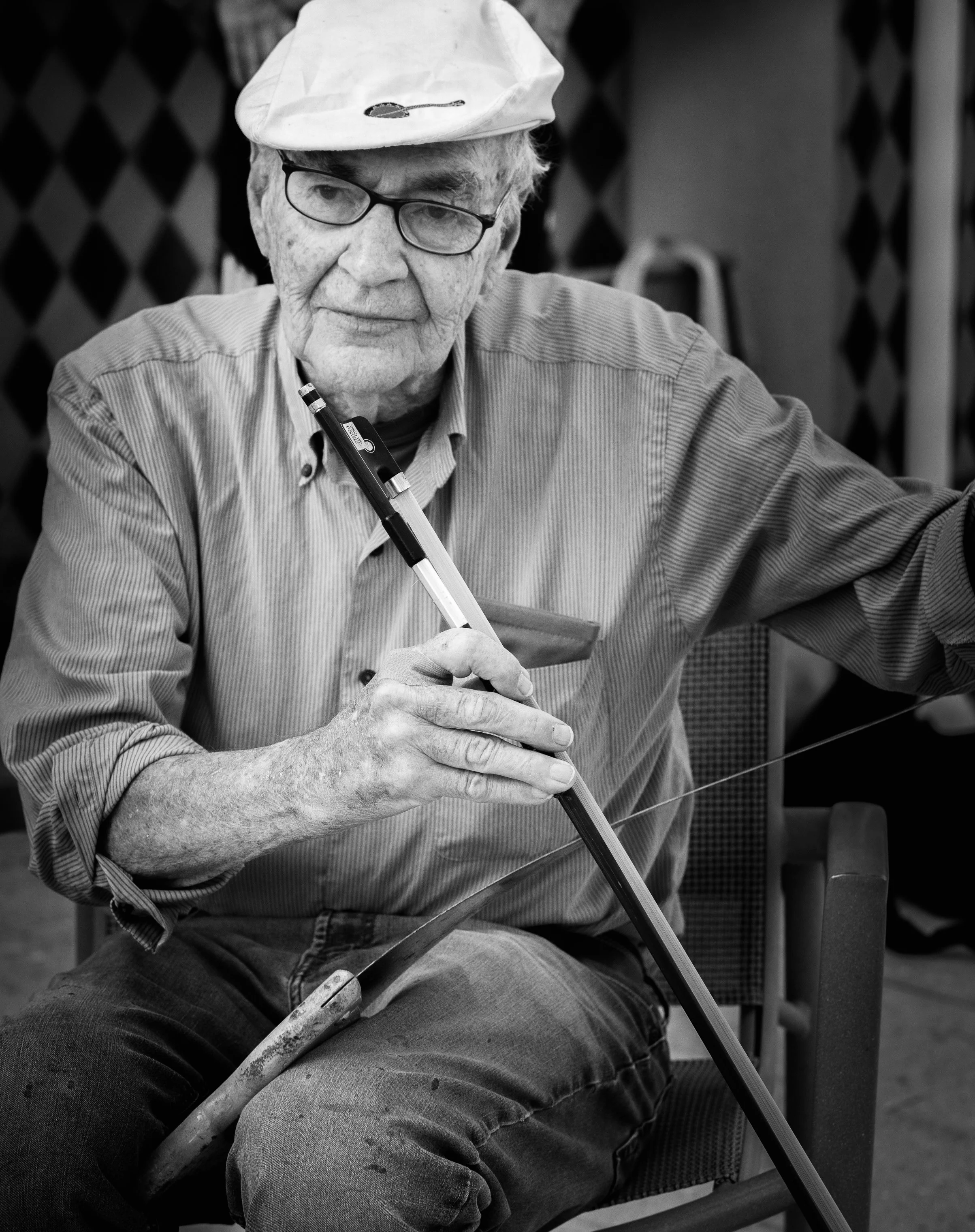 Older man with glasses, wearing a cap, sitting and writing with a pen in his right hand, with a background of a patterned wall.