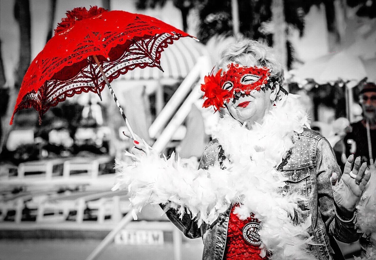 A woman dressed in a red costume with a red feathered mask and holding a red parasol, with the rest of the image in black and white.