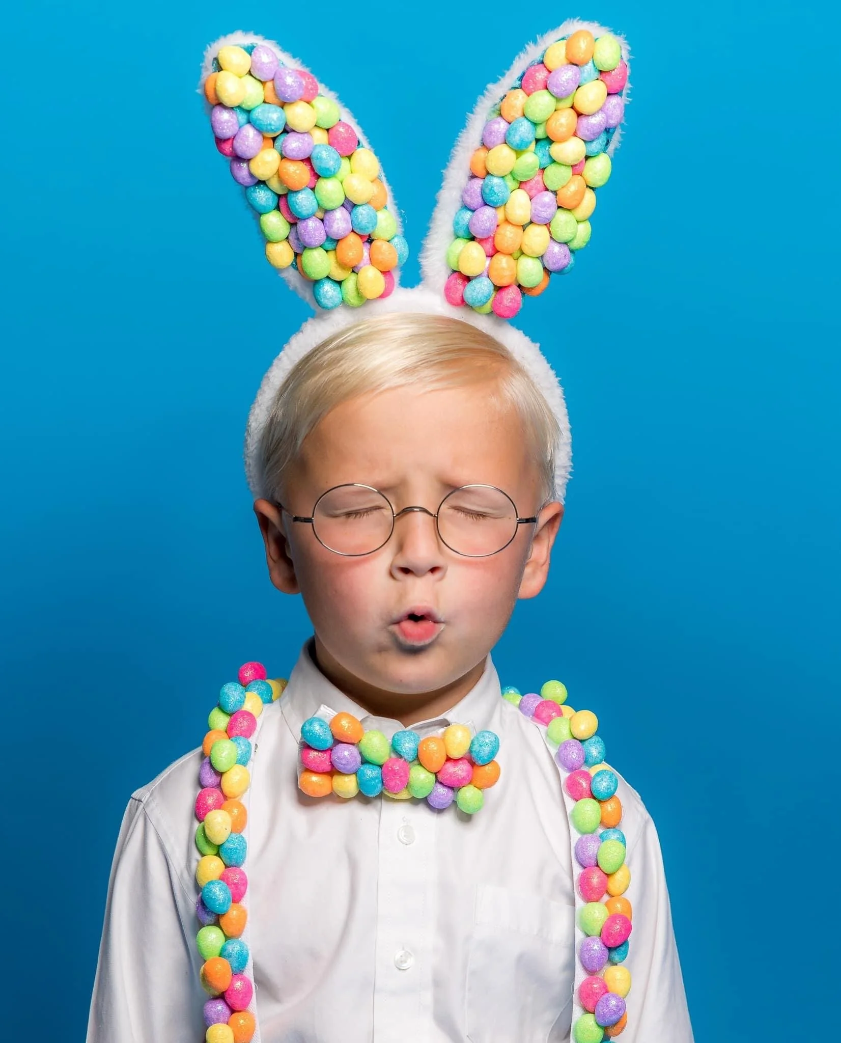 A person with blond hair, glasses, and dressed in a white shirt, wearing colorful Easter bunny ears and a matching necklace made of pastel-colored plastic eggs, against a bright blue background.