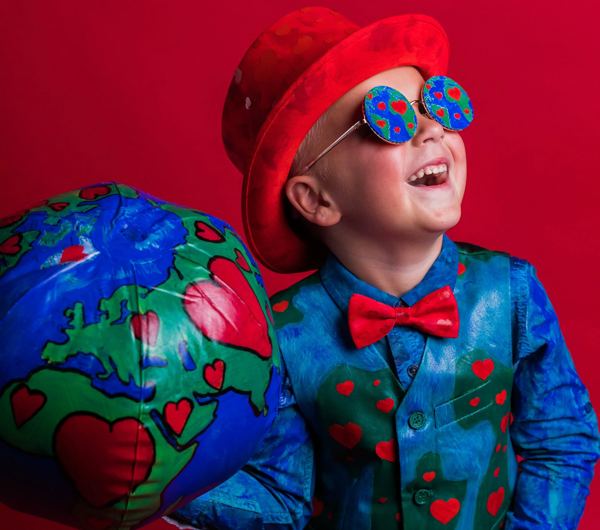 A young child in a red hat and heart-patterned clothing wearing heart-themed sunglasses, smiling with mouth open against a red background.