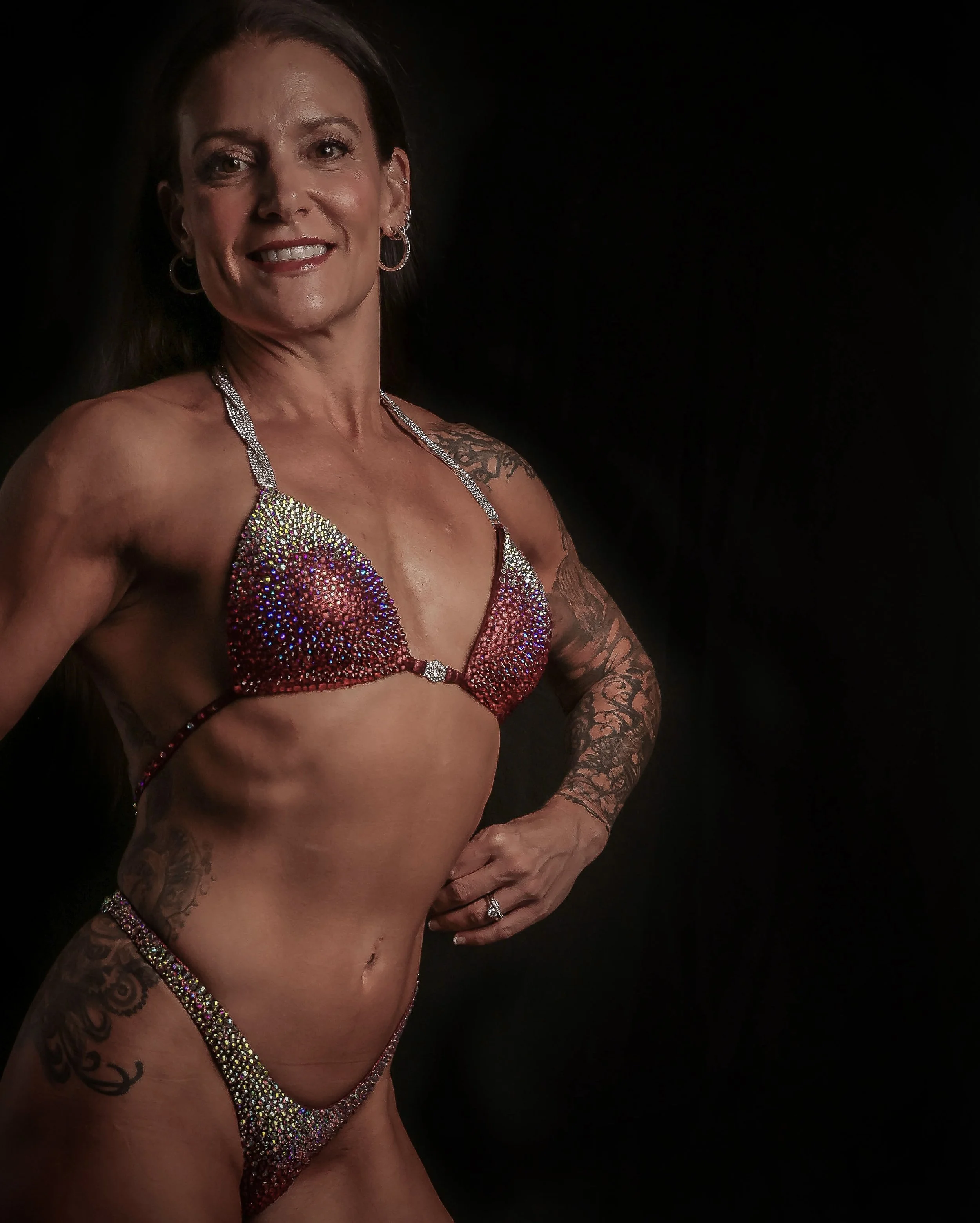 A woman with dark hair and tattoos wearing a bejeweled bikini posing against a black background.