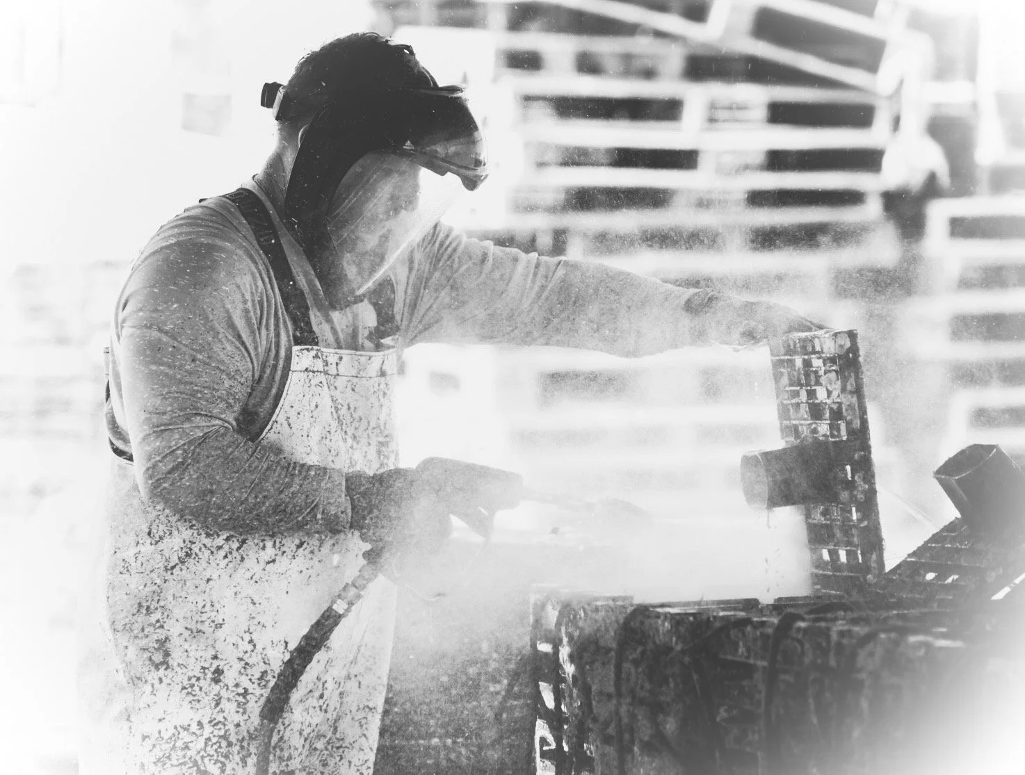 A person wearing a protective face shield, a long-sleeve shirt, and a work apron is using a power tool on a piece of machinery, creating dust or sparks in a workshop or industrial setting.