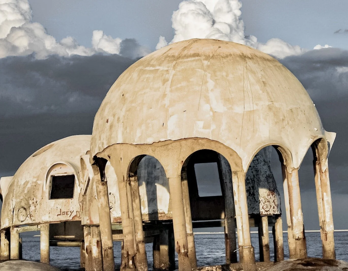 Abandoned, deteriorated concrete domed structure with multiple arches, located near the water, under a cloudy sky.