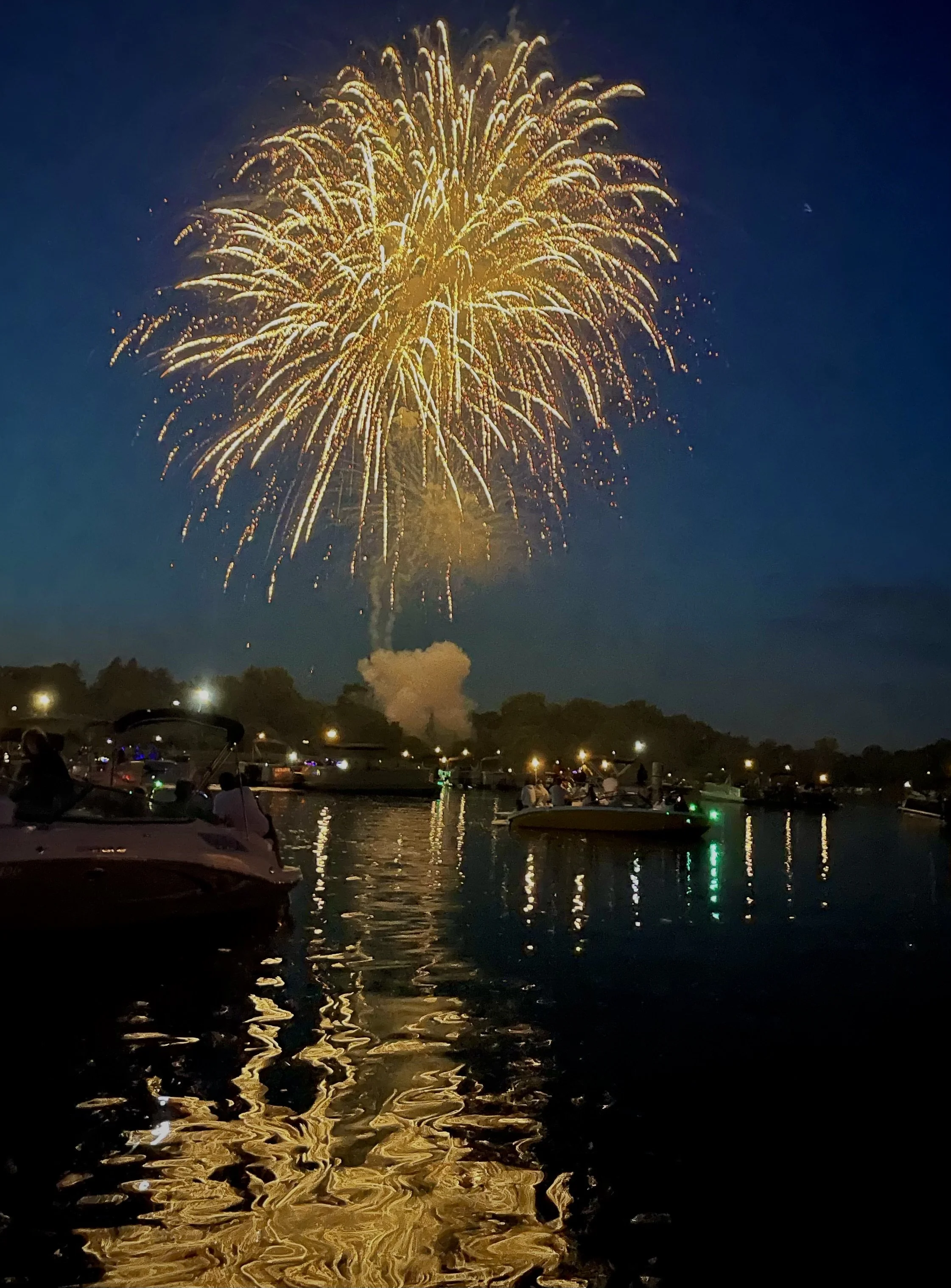 Fireworks exploding in a night sky over a marina with boats reflecting the golden and white light on the water.