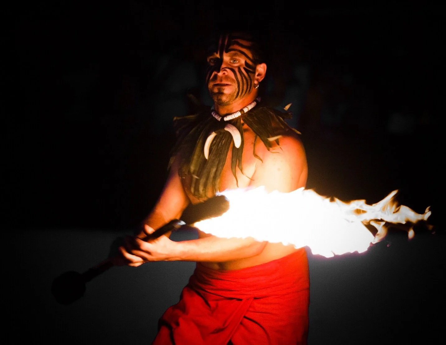 A man performing fire dance at night, holding a flaming torch, with tribal face paint, a necklace of bones, and wearing red clothing.