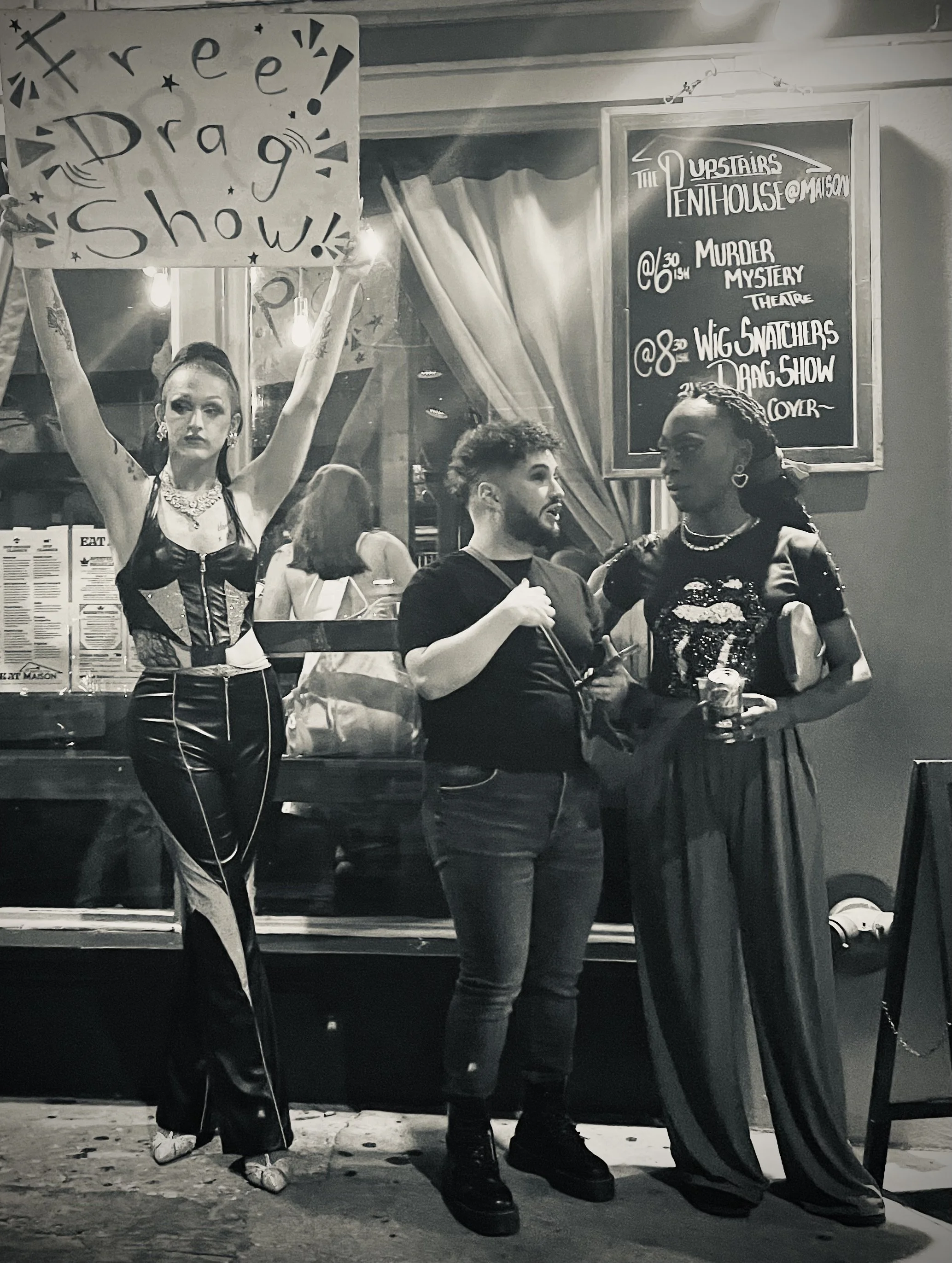 People at a social event. One woman holds a large sign that says 'Free Drag Show!' The image is in black and white.