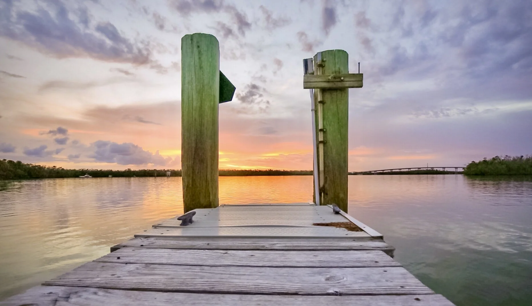 A wooden dock extends over calm water during sunset with a colorful sky of orange, pink, and purple hues, and a distant bridge in the background.