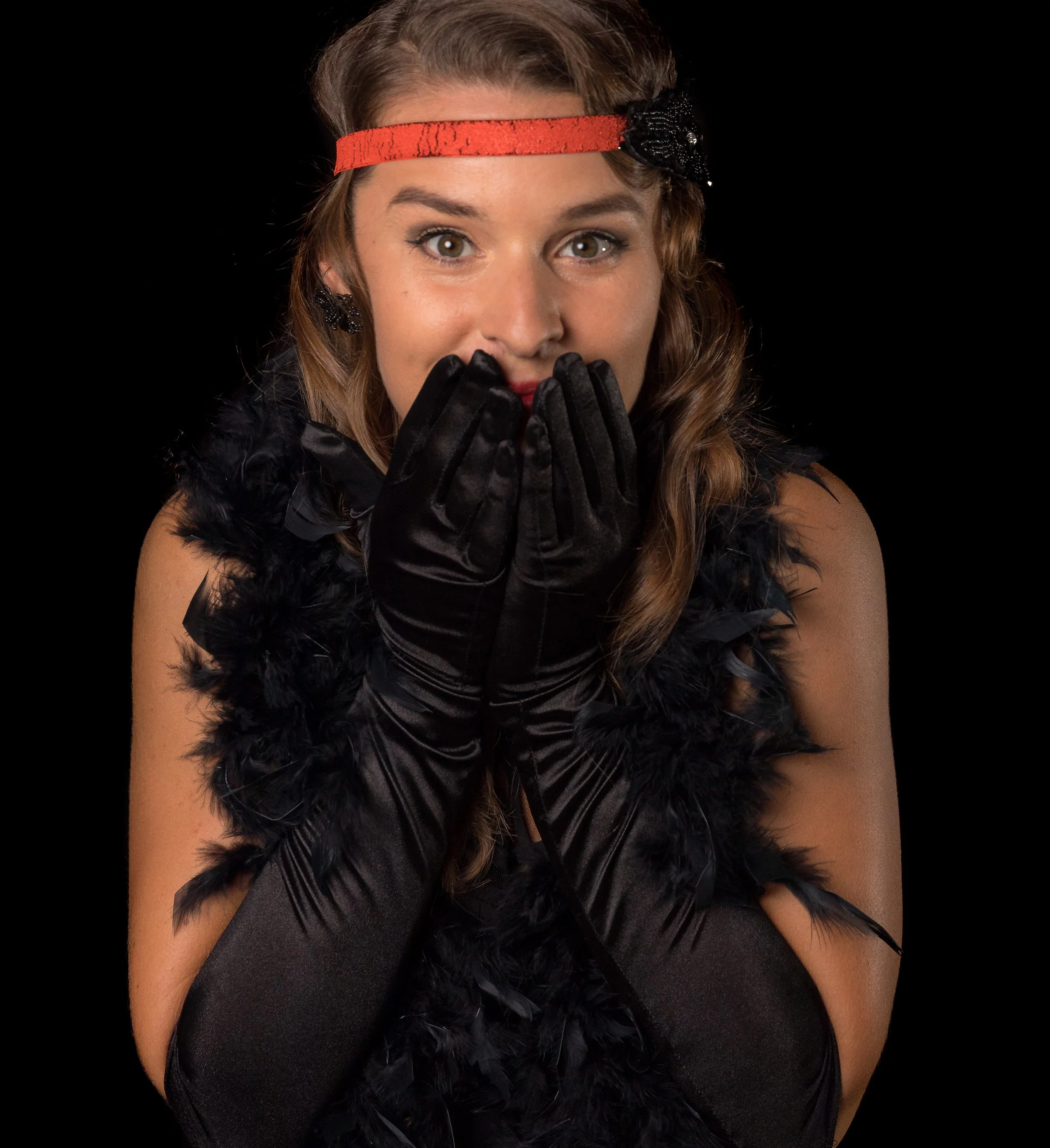 Close-up of a woman with light brown hair and brown eyes, wearing a red headband with a black embellishment, black feathered garment, black gloves, and dark top, holding her hands near her mouth with a surprised or playful expression.