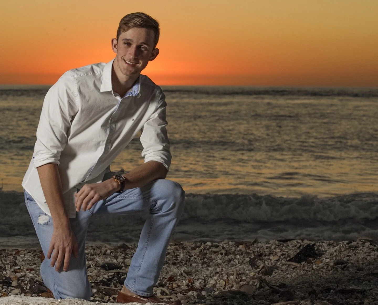 A young man in a white shirt and ripped jeans squatting on a rocky beach at sunset, smiling at the camera with the ocean and orange sky in the background.