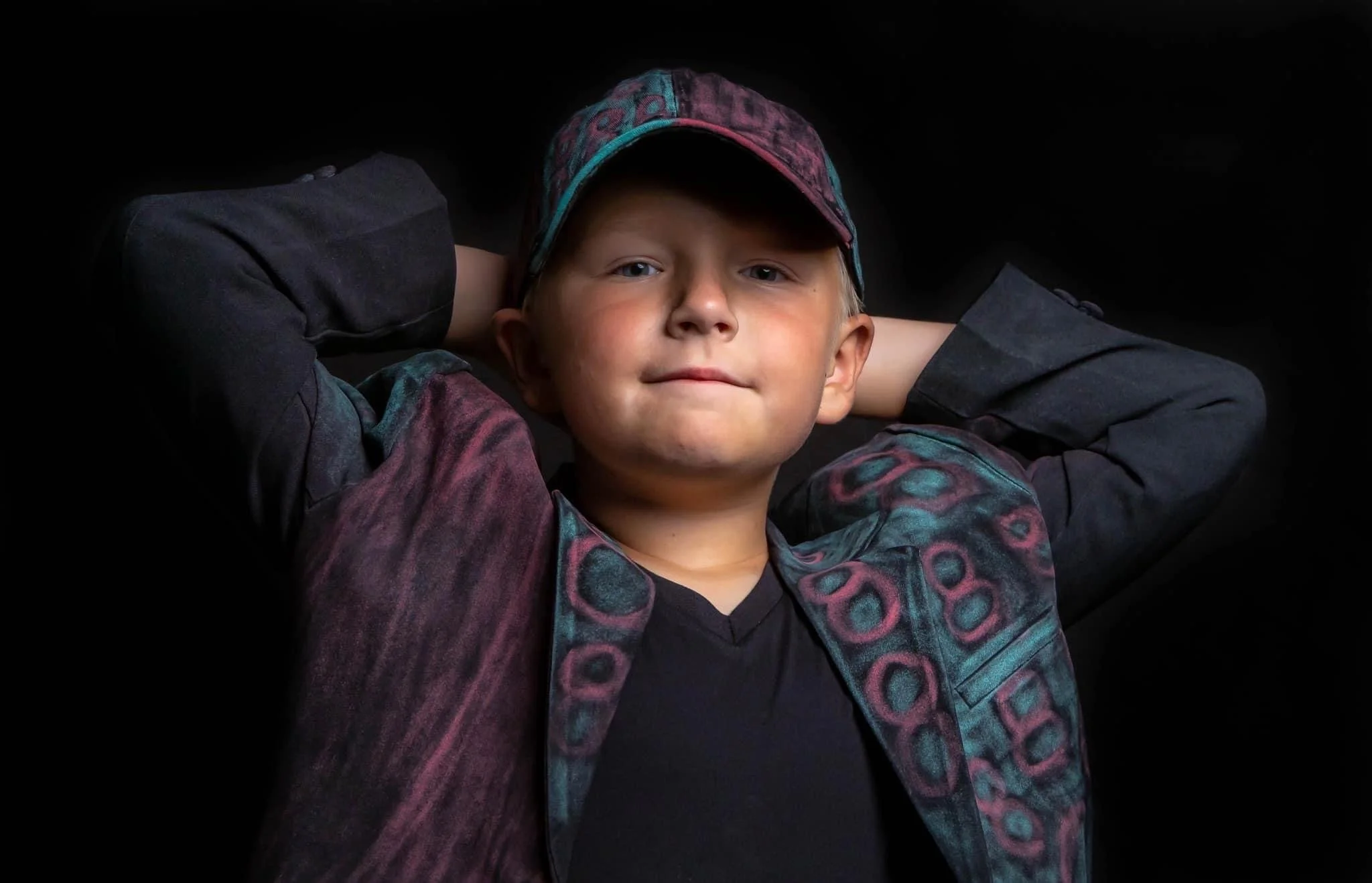 A young boy with blond hair, wearing a colorful jacket and baseball cap, lying back with hands behind his head against a black background.