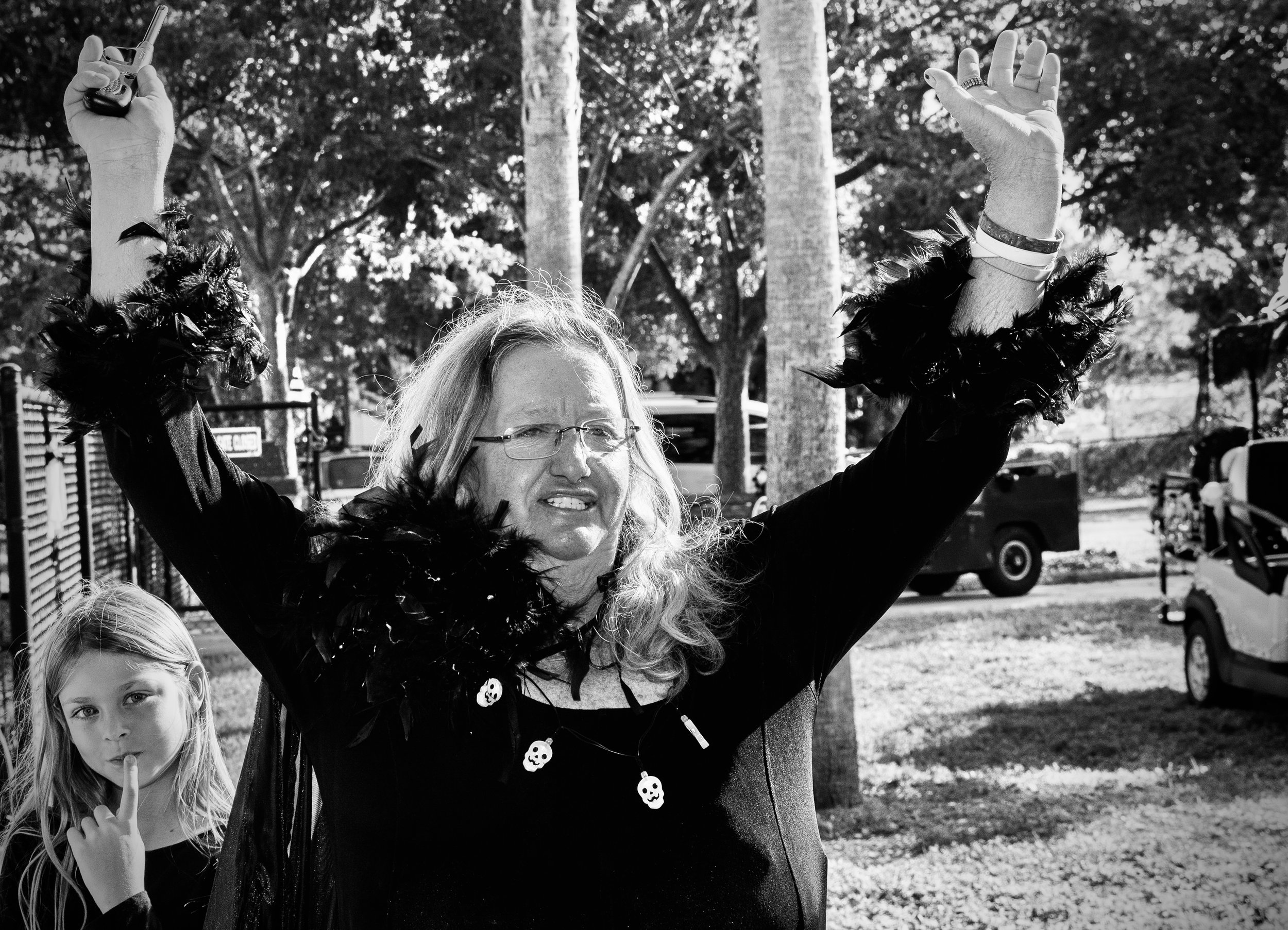 A woman and a young girl outdoors in black and white. The woman is holding her arms up, wearing festive accessories, and the girl is standing next to her, also making a gesture with her finger to her lips.