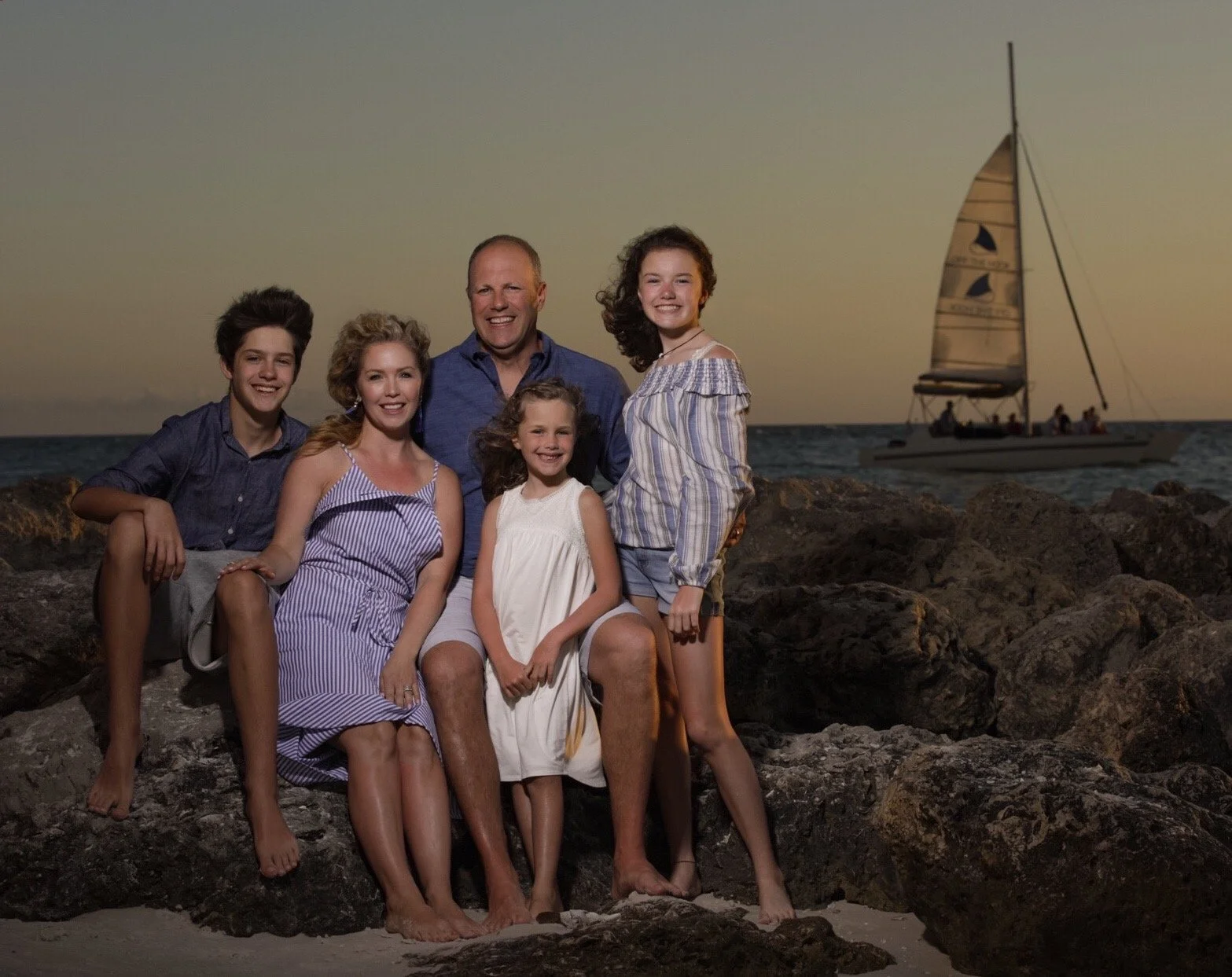 Family of six posing on rocks at the beach during sunset, with sailboat in background.