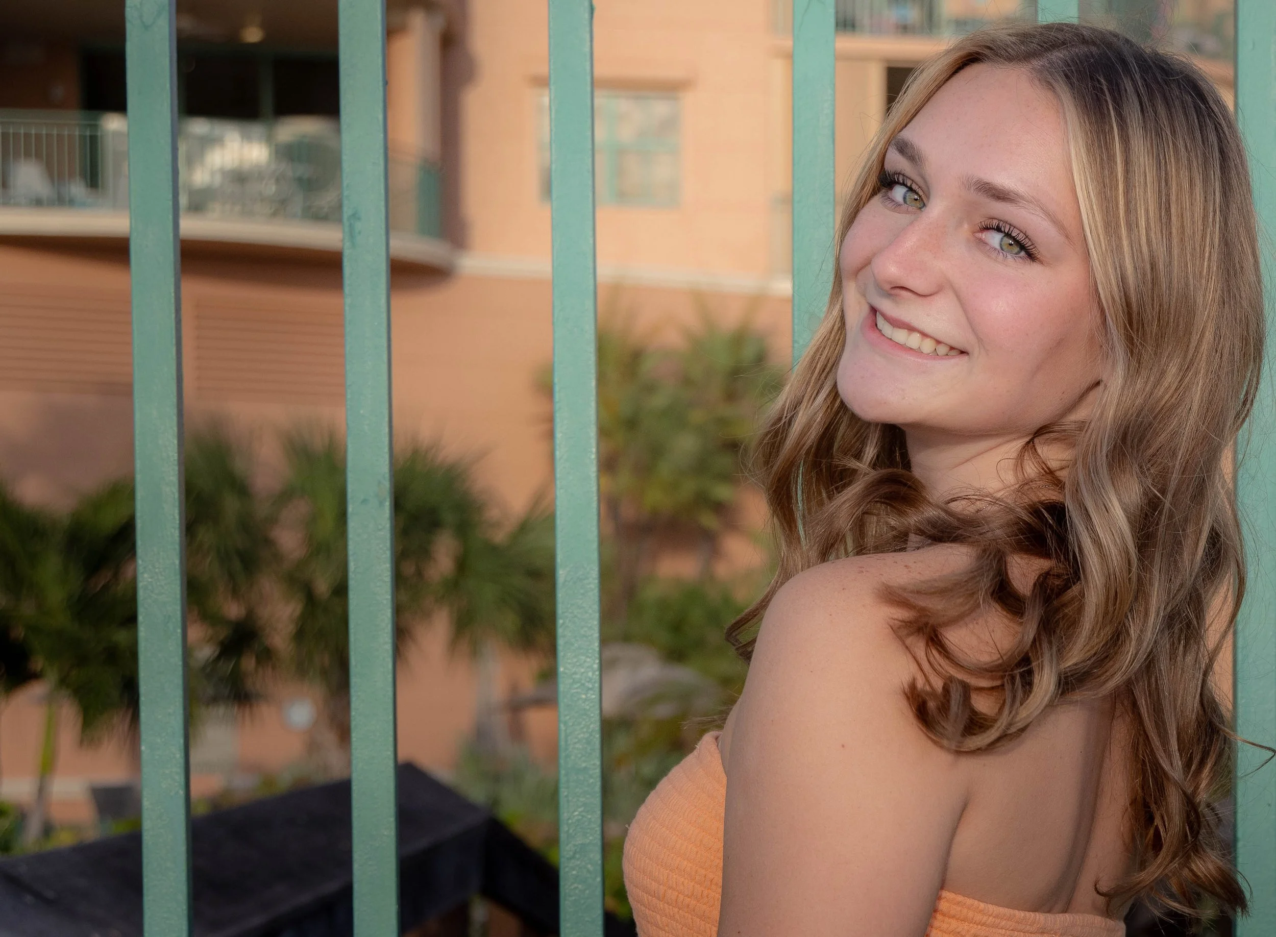 A young woman with long, wavy, light brown hair and light-colored eyes, smiling and leaning against a green metal fence, with a blurred background of a residential building and greenery.