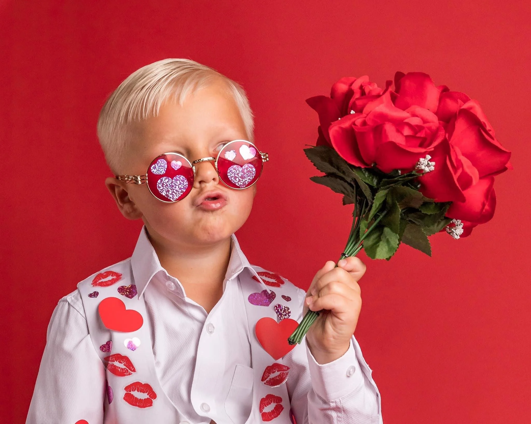 Child with blond hair wearing heart-shaped sunglasses with love-themed designs and a white shirt decorated with heart and kiss marks, holding a bouquet of red roses against a red background.