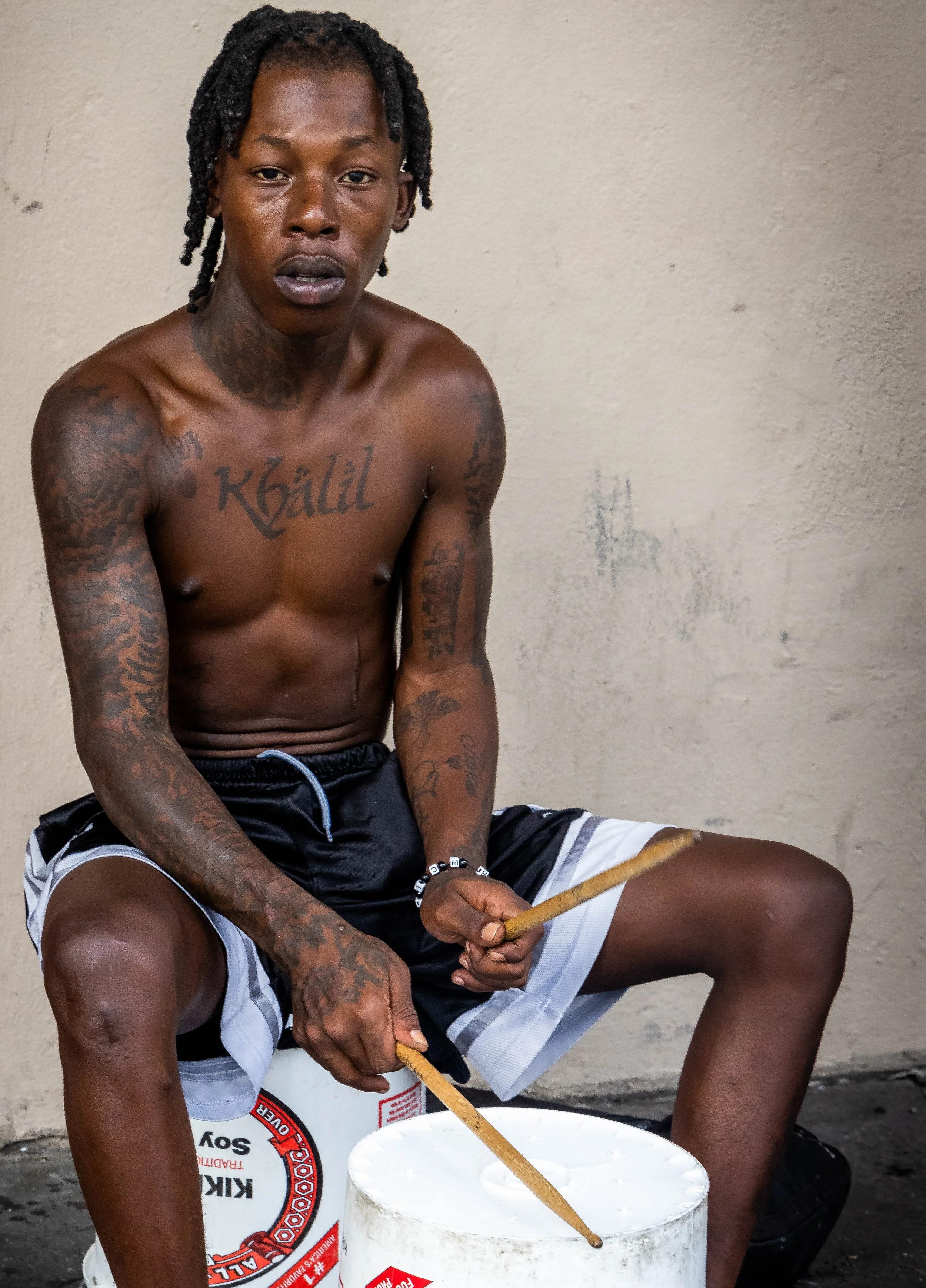 A shirtless man with tattoos on his arms and chest, sitting against a plain wall, holding two wooden sticks over a plastic bucket of liquid, with a slightly open mouth and a serious expression.