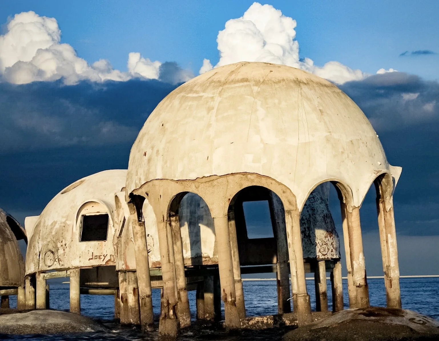 Abandoned white domed building with arches and columns, partly submerged in water under a cloudy sky.