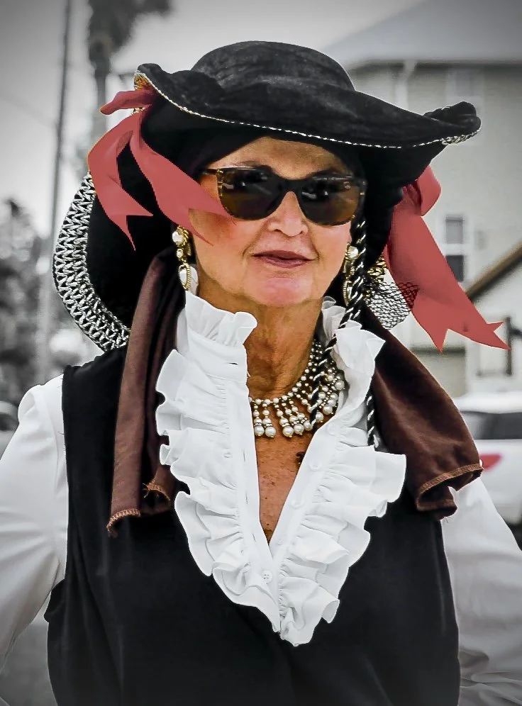 An older woman dressed in vintage style with a black wide-brimmed hat adorned with red ribbons, dark sunglasses, pearl necklace, and pearl earrings. She is wearing a white blouse with ruffled front and a black vest. The background is outdoors with ho